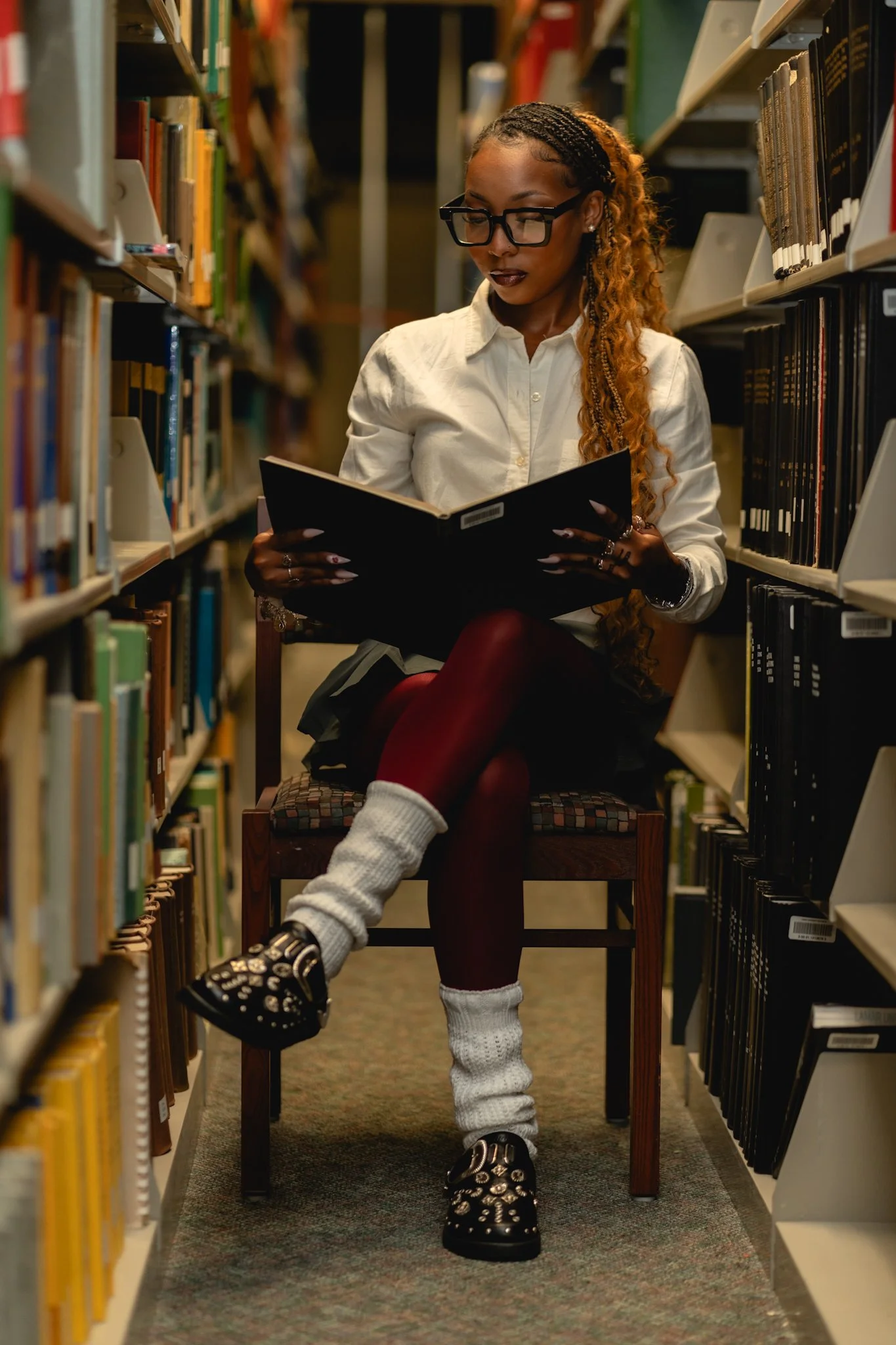 A person with glasses and braided hair is sitting on a chair in a library, reading a book. They are wearing a white shirt, maroon leggings, and black shoes with white socks. Bookshelves filled with colorful books are on either side.