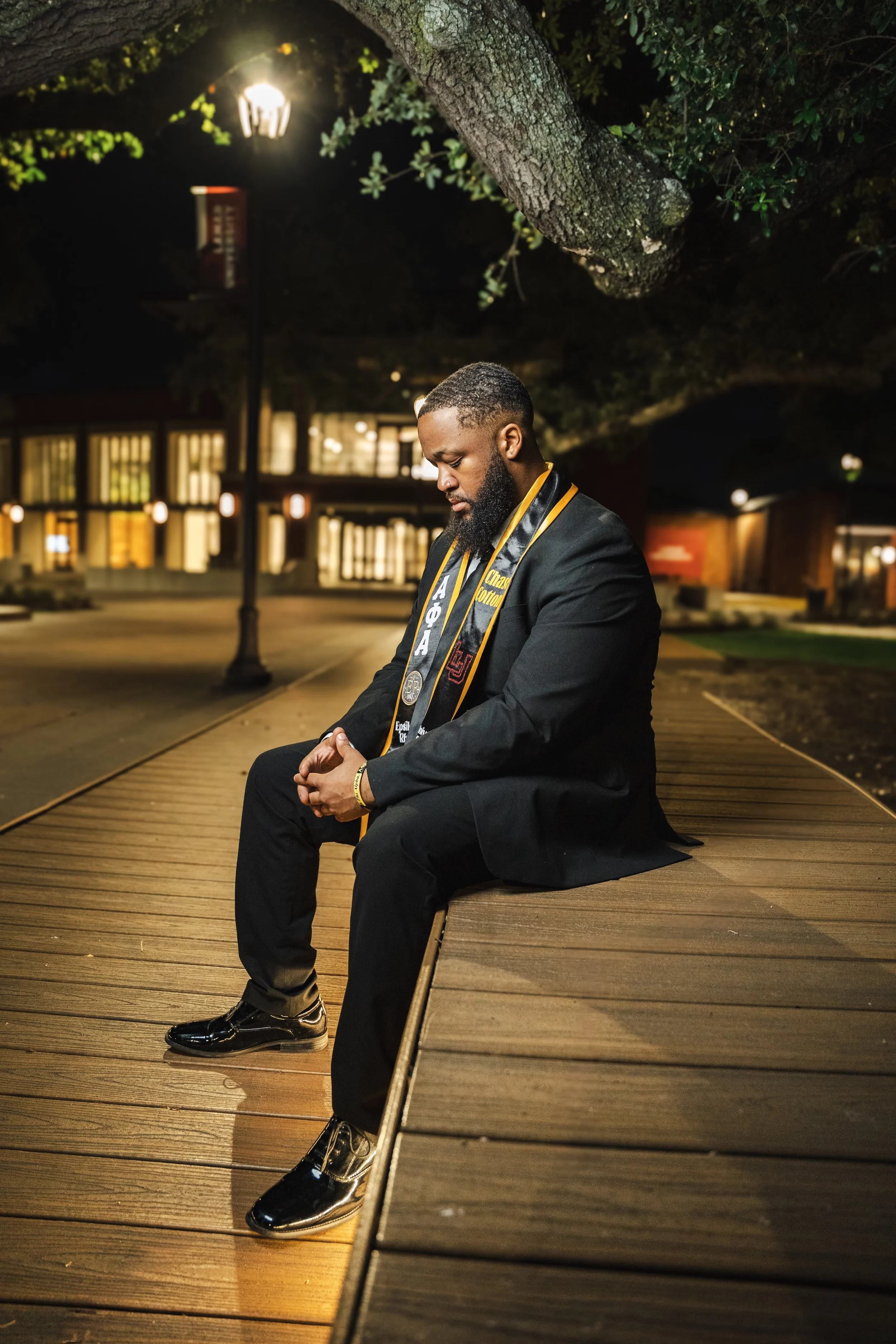 A man in a formal suit with a graduation stole sits on a wooden path at night, illuminated by a street lamp, with trees and a building in the background.