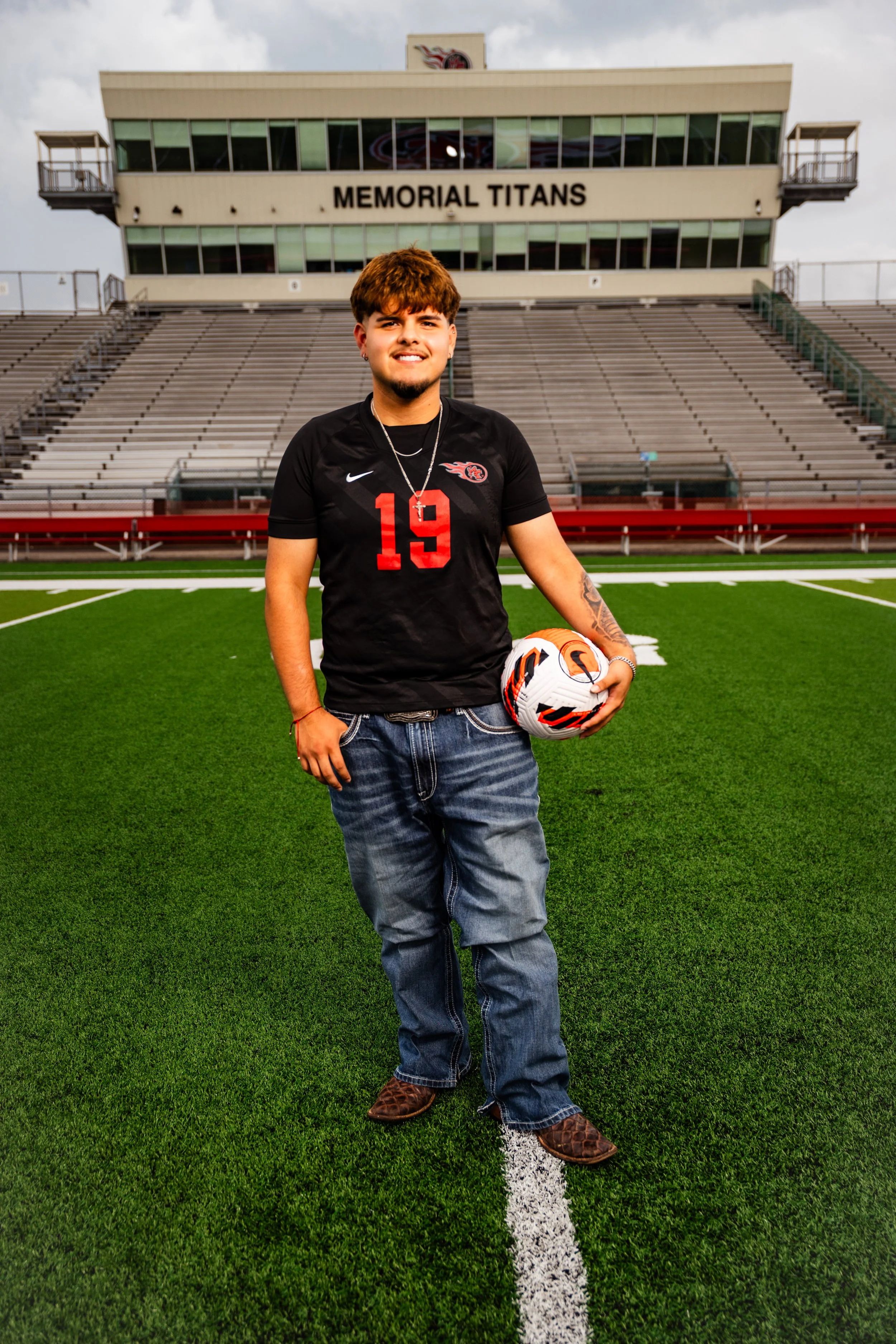 A young man in a black sports jersey and jeans standing on a football field, holding a soccer ball, with stadium stands labeled "Memorial Titans" in the background.