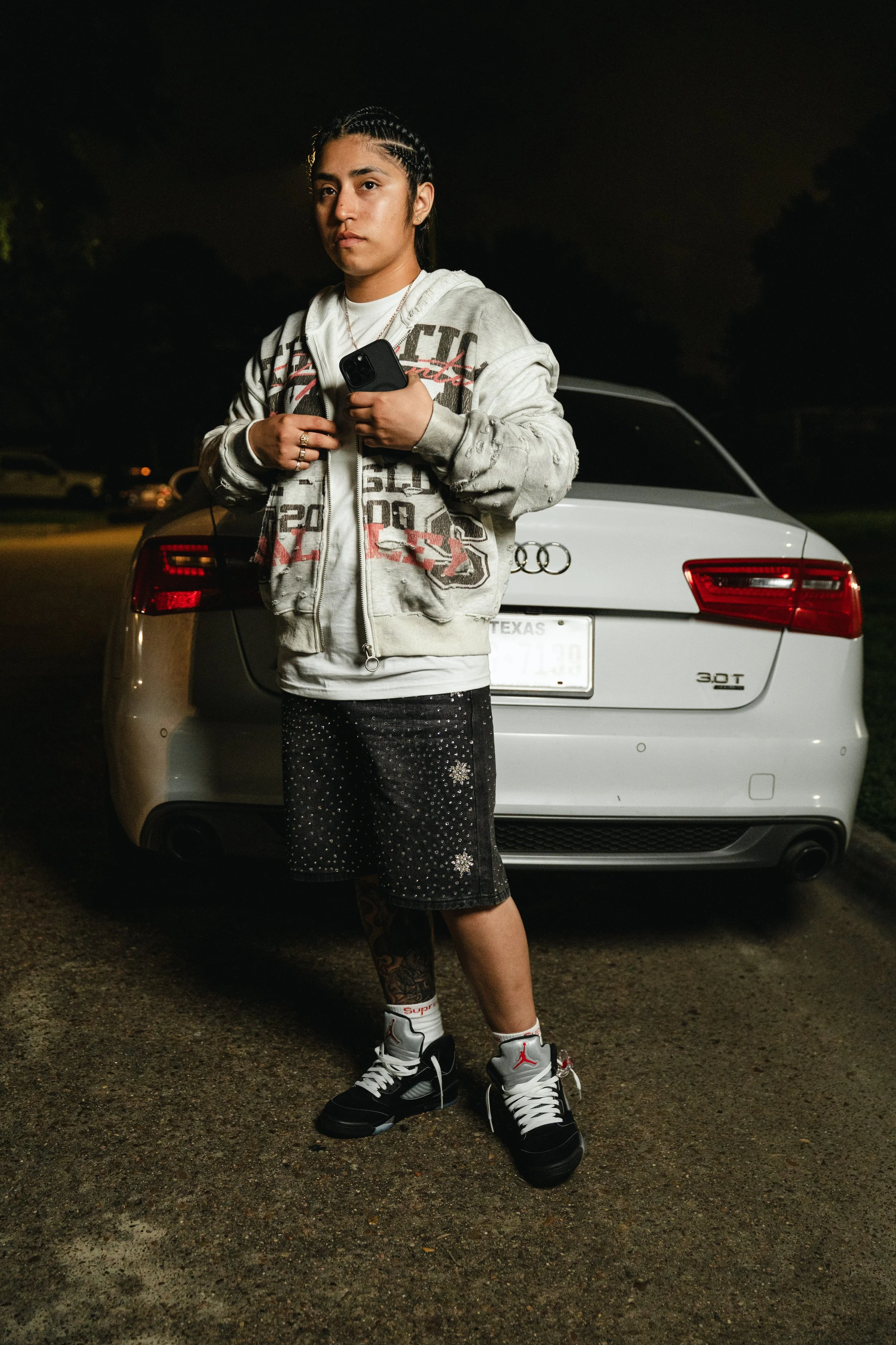 Person standing in front of a white Audi car at night, wearing a graphic hoodie, black shorts, and sneakers.