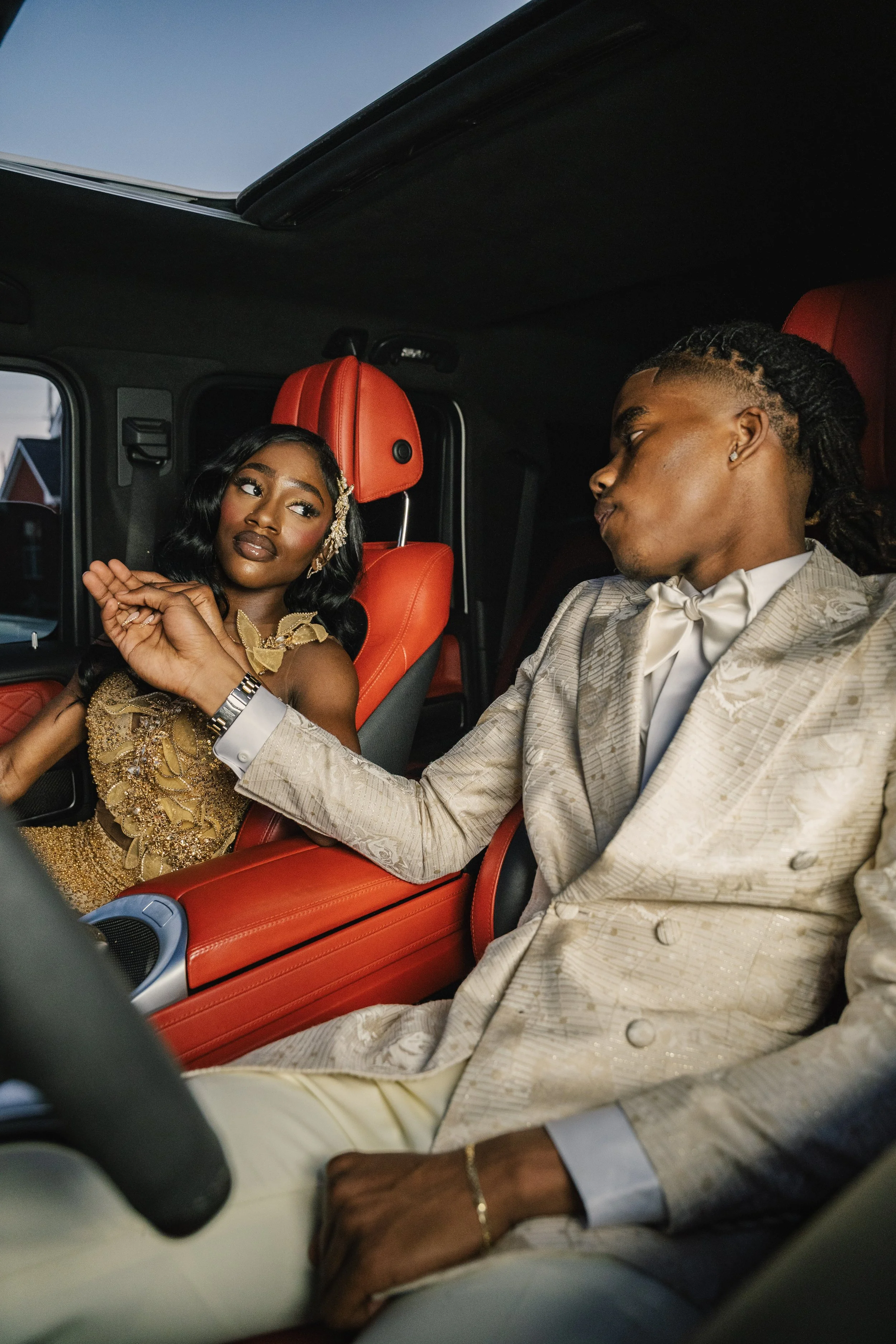 A couple dressed in elegant attire, sitting in a vehicle with red leather seats. The woman is wearing a gold dress and has styled hair. The man is wearing a light-colored suit with a bow tie. They are holding hands and sitting in the front seats of the car.