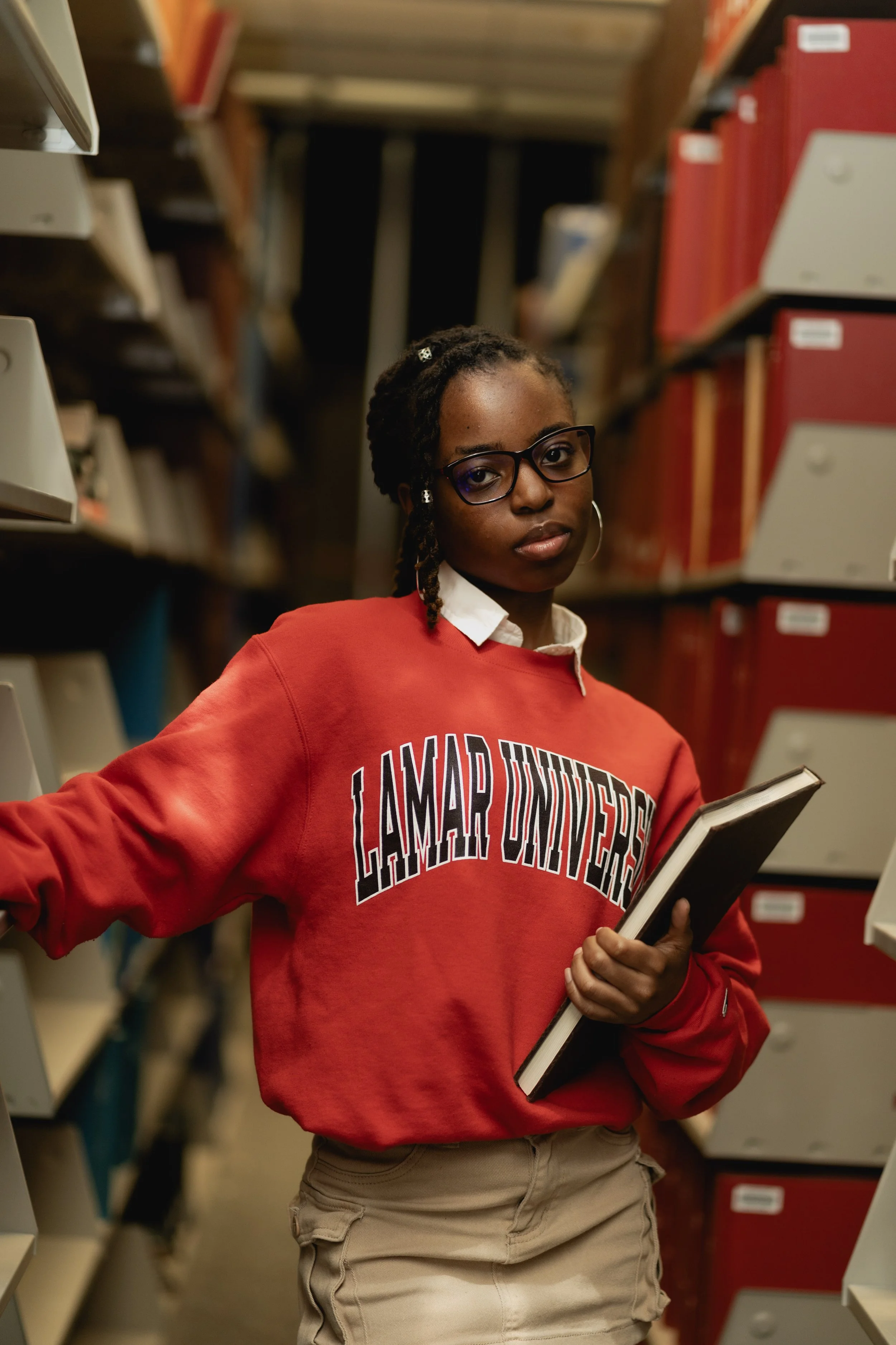 A person wearing glasses and a "Lamar University" red sweatshirt holds a book while standing in a library aisle.