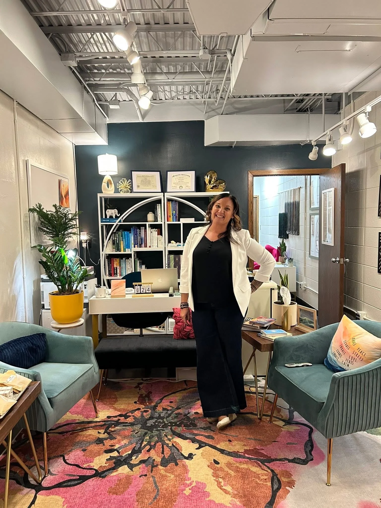 A woman with dark hair and light skin wearing a white blazer and black dress smiling in a modern, cozy room filled with books, plants, and colorful decor.