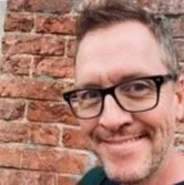 Man with glasses standing in front of a brick wall