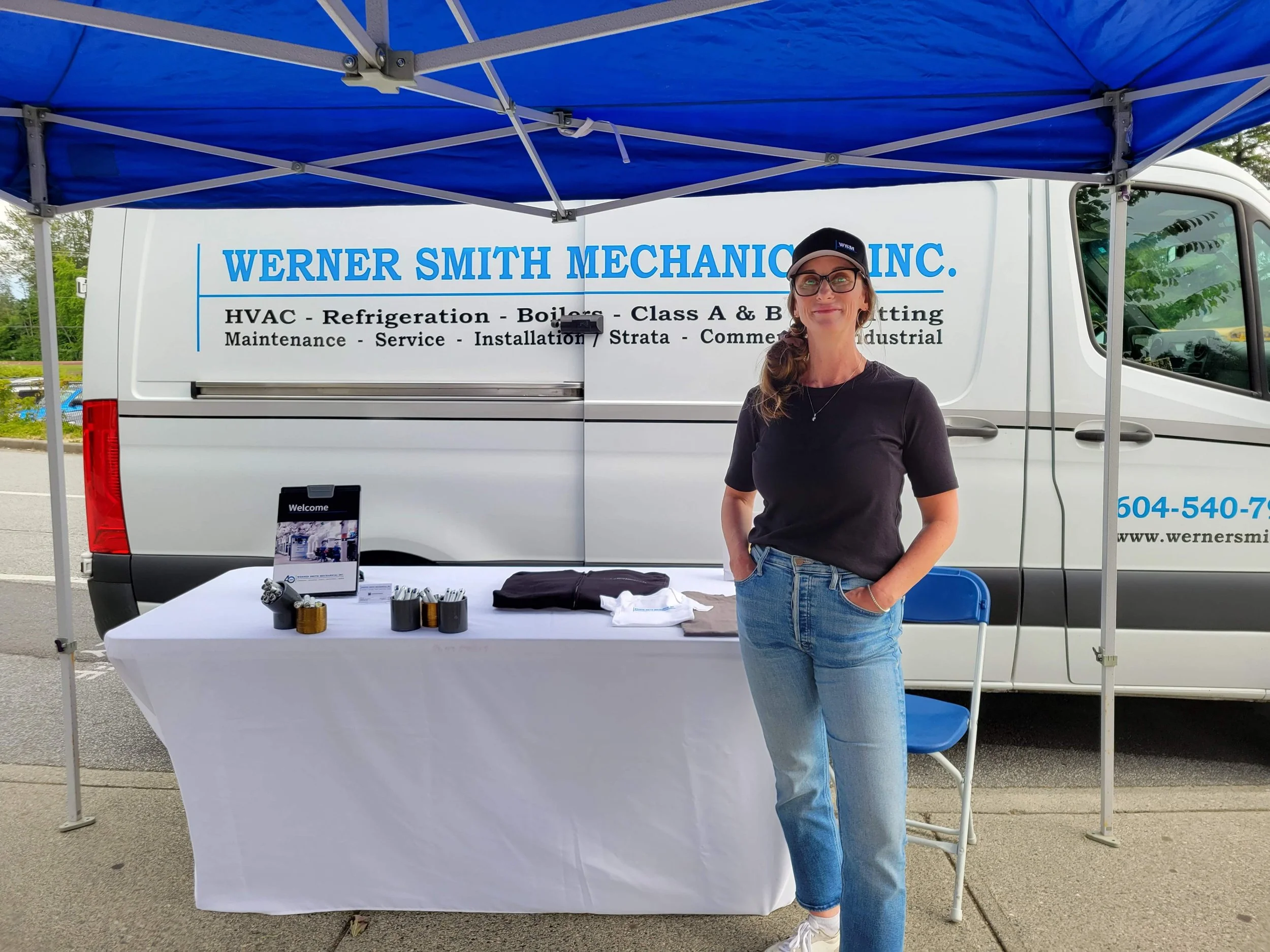 a woman in a baseball hat, jeans and a t-shirt, standing outside in front of a company work van and a table at a career fair