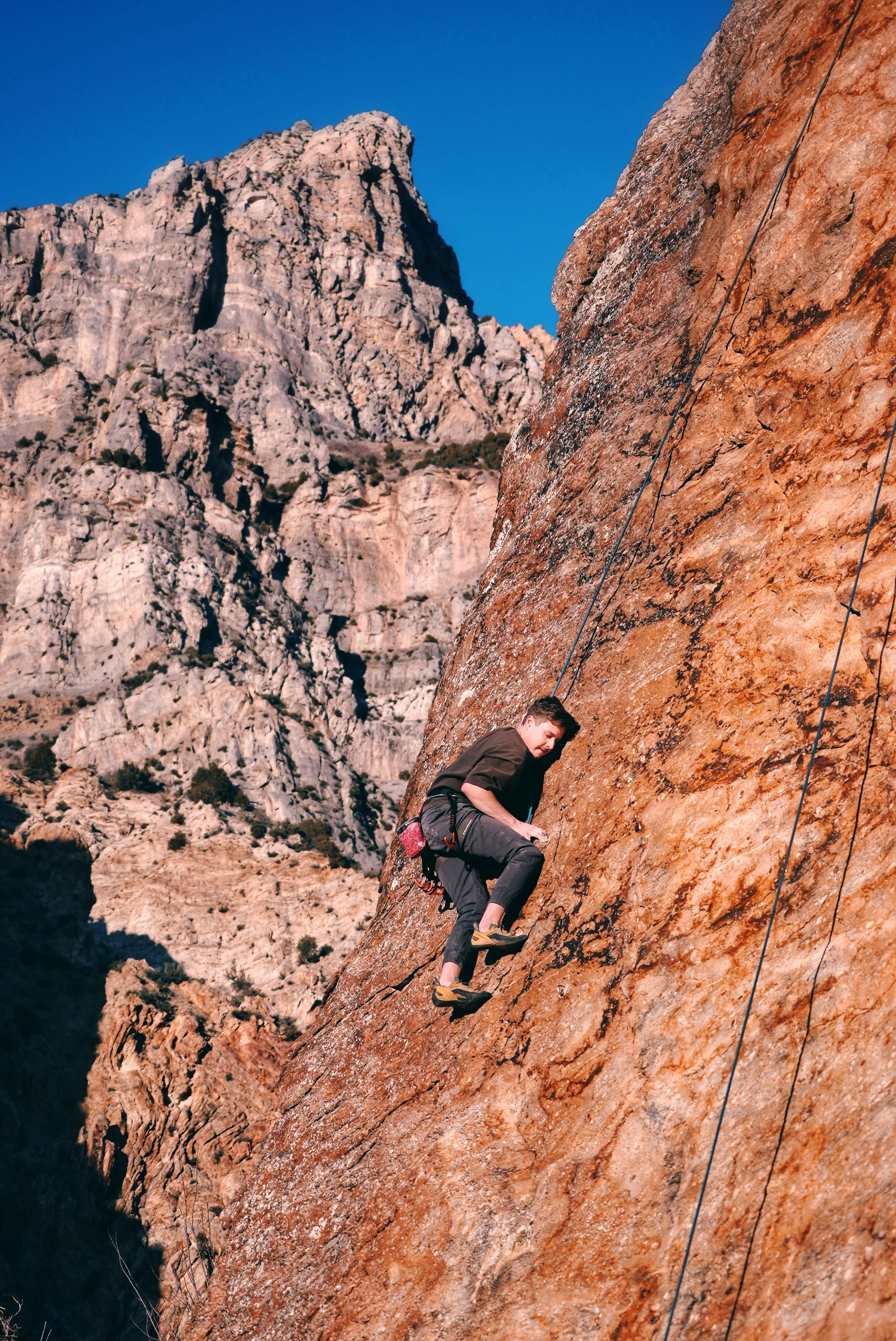 A person rock climbing on a steep, reddish-brown cliff face with mountains under a clear blue sky in the background.