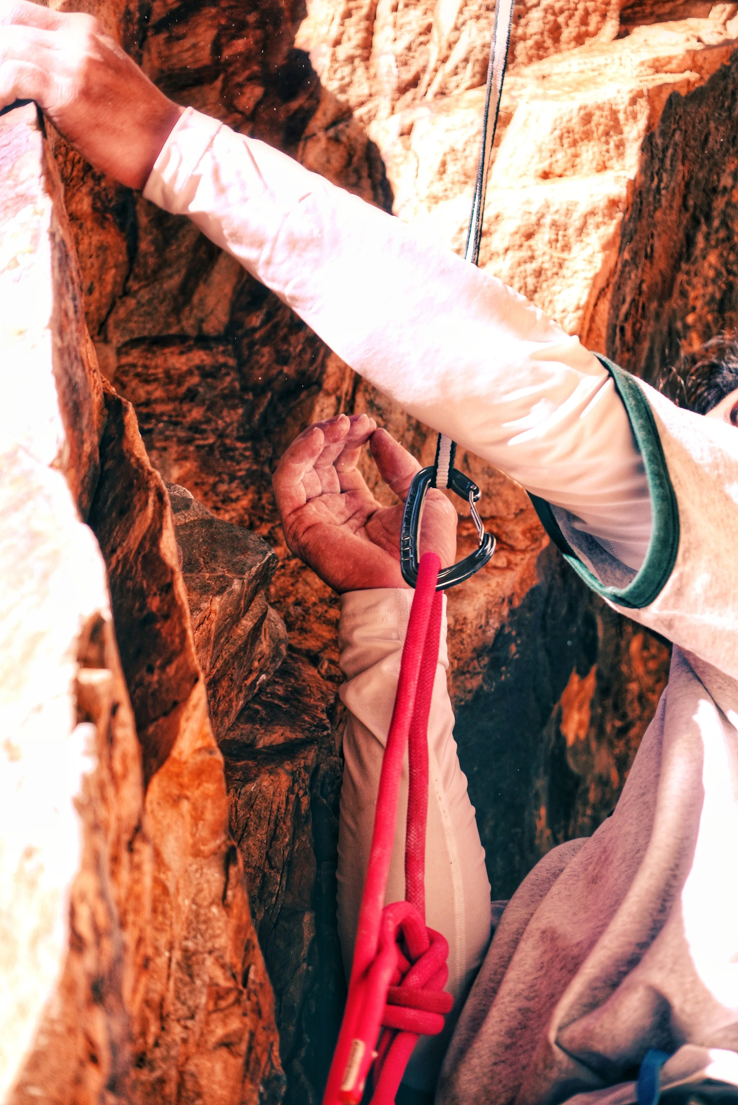 Climber gripping rock with hand, wearing a white jacket, carabiner attached to harness, red climbing rope secured on rock, in a narrow canyon.