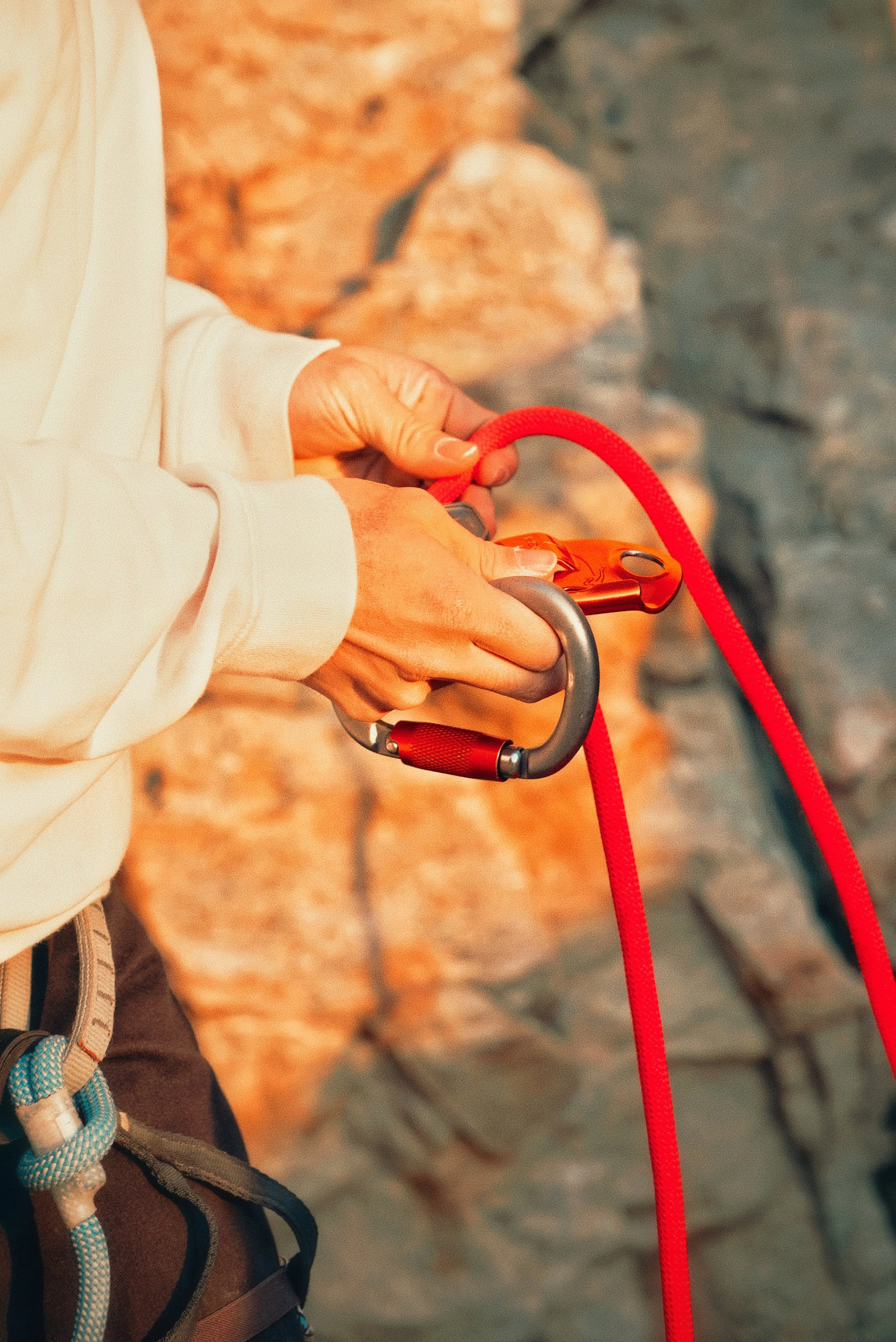 Close-up of a person's hands gripping a red and black climbing carabiner attached to a red climbing rope, set against a rocky background.