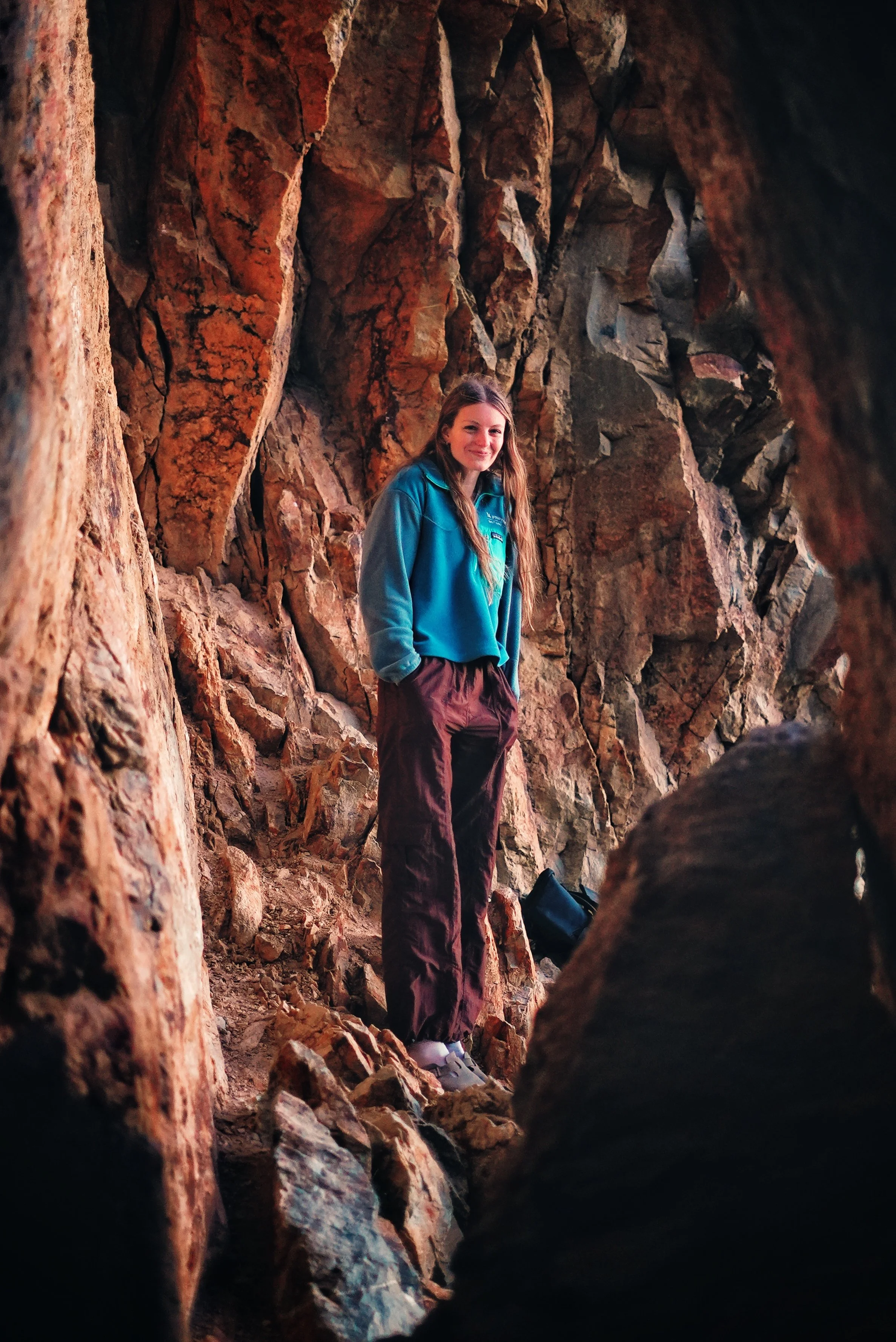 A young woman standing inside a rocky canyon or crevice, smiling, wearing a blue jacket and brown pants, with a backpack on the ground nearby.