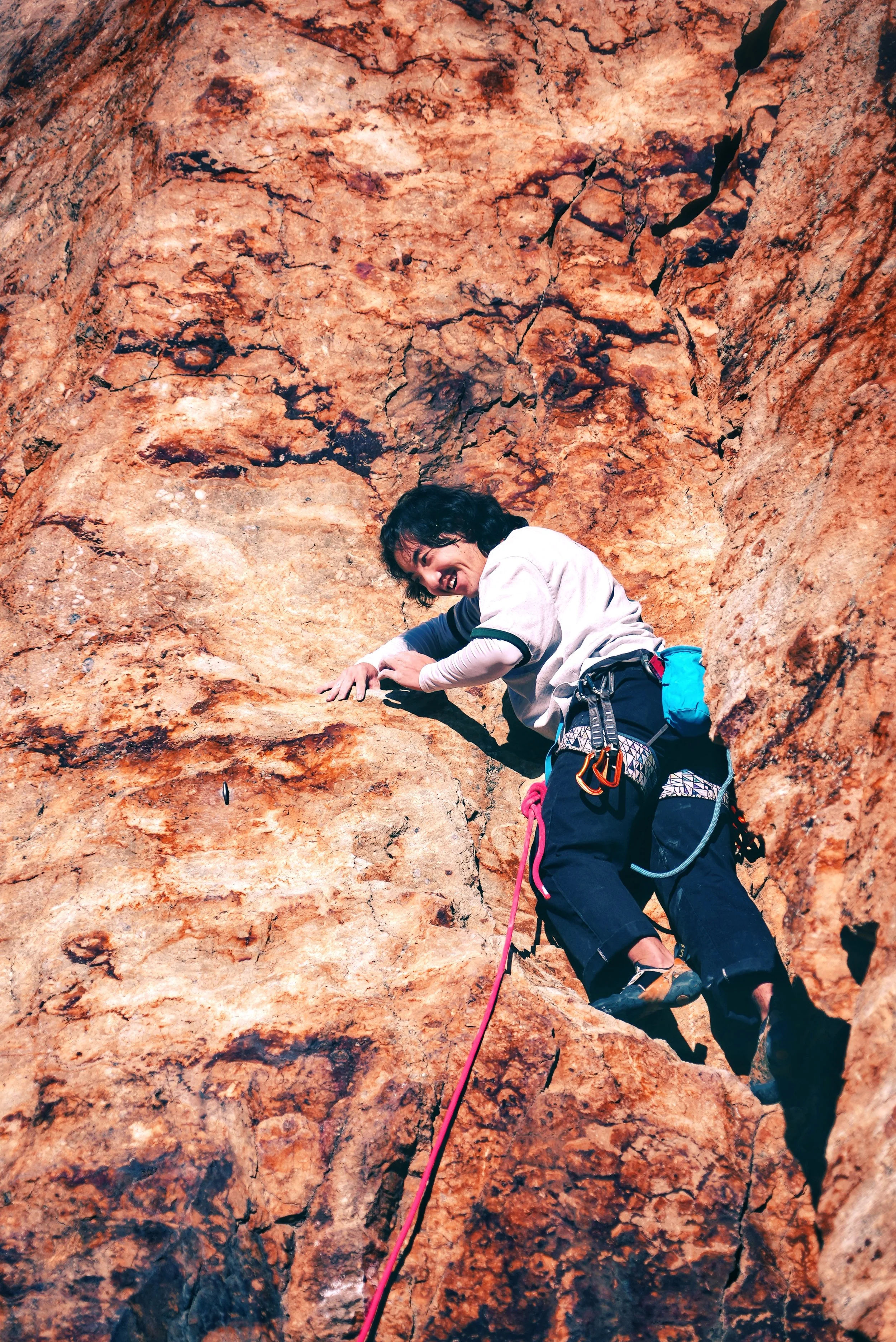 A woman rock climbing on a steep, reddish-brown cliff, smiling and holding onto the rocks with climbing gear attached to her harness.