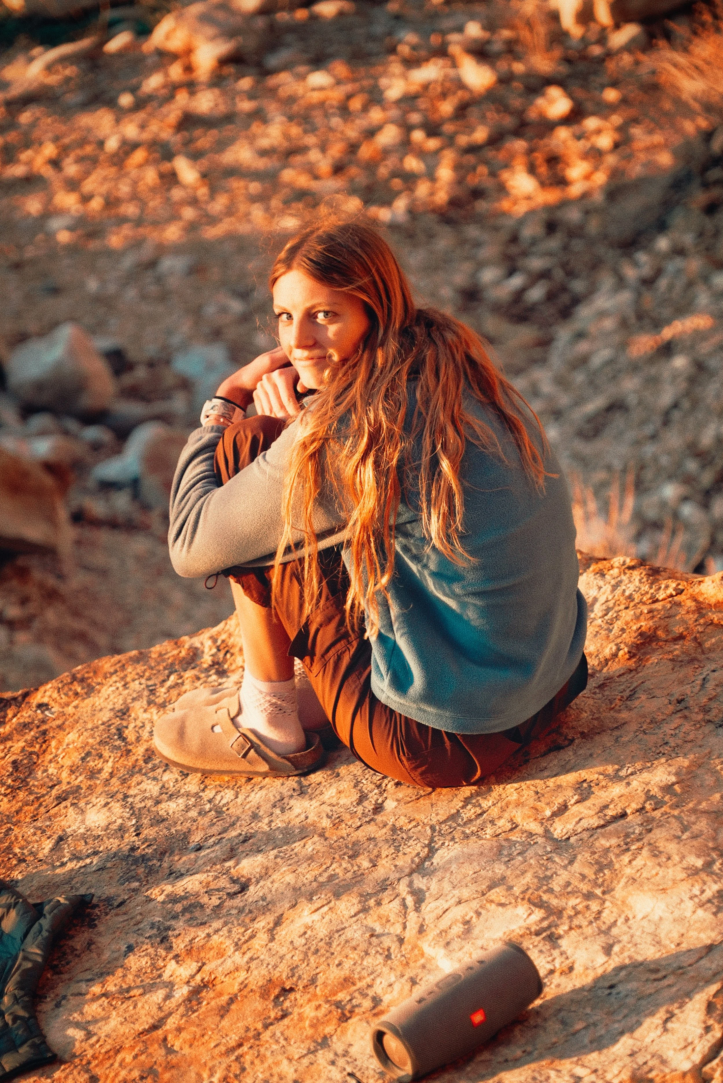 Young woman sitting on a rock in a rocky outdoor area at sunset, with a portable speaker nearby.