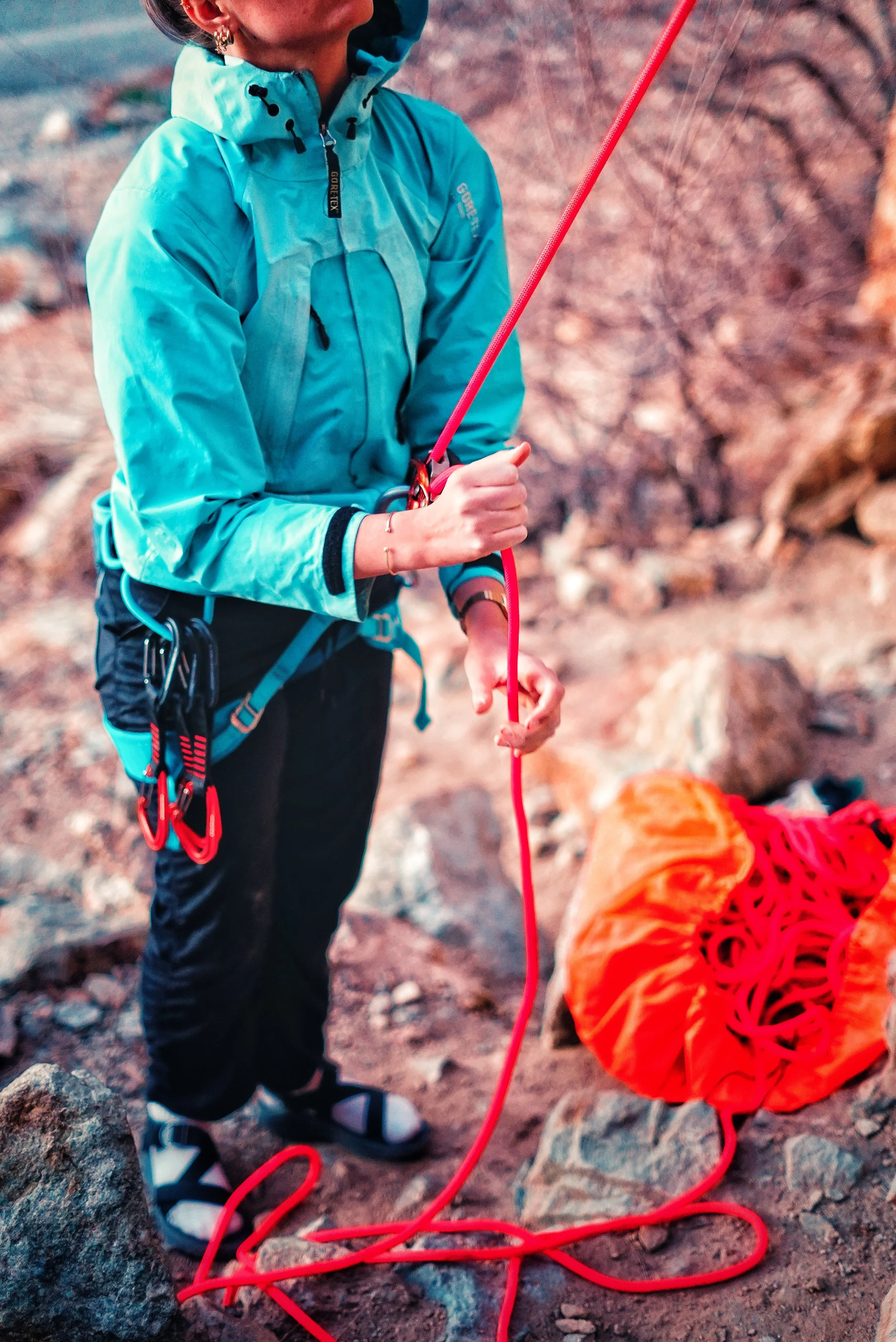 A woman in a turquoise outdoor jacket and black pants is preparing climbing gear, with a coiled orange rope in an orange bag on rocky terrain.