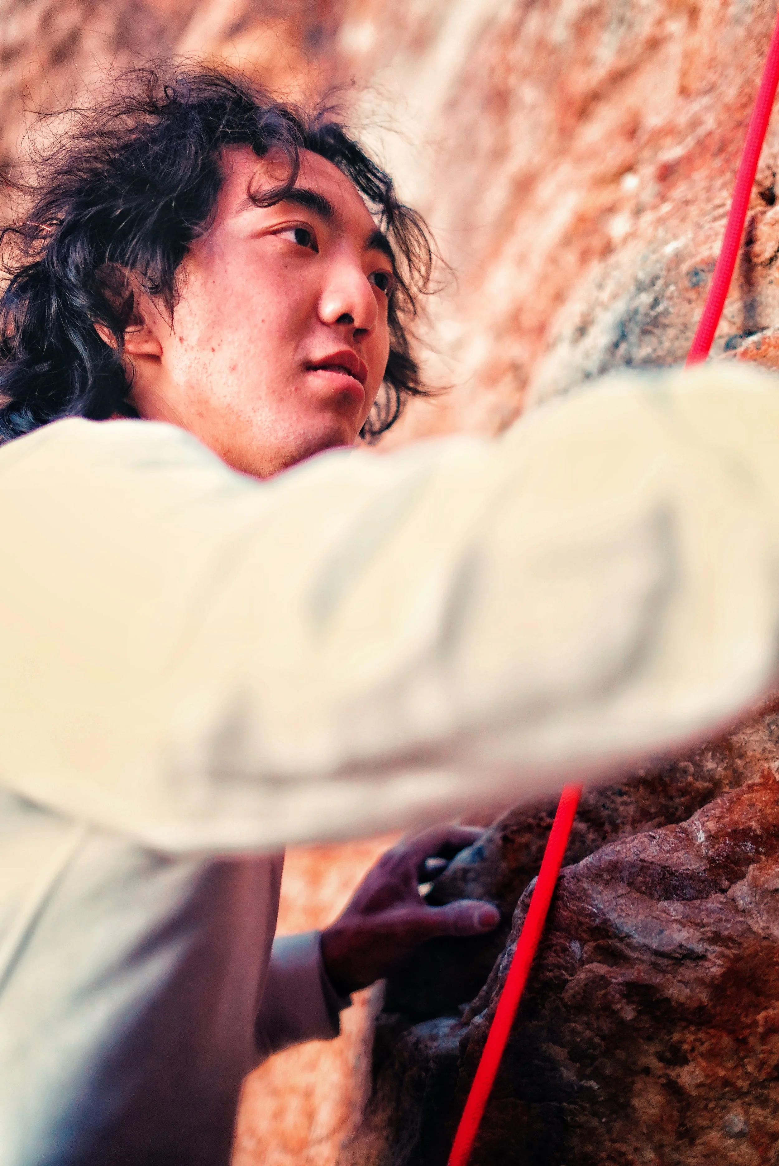 A person with curly hair rock climbing, reaching for a hold on an indoor or outdoor rock wall, with a red climbing rope visible.