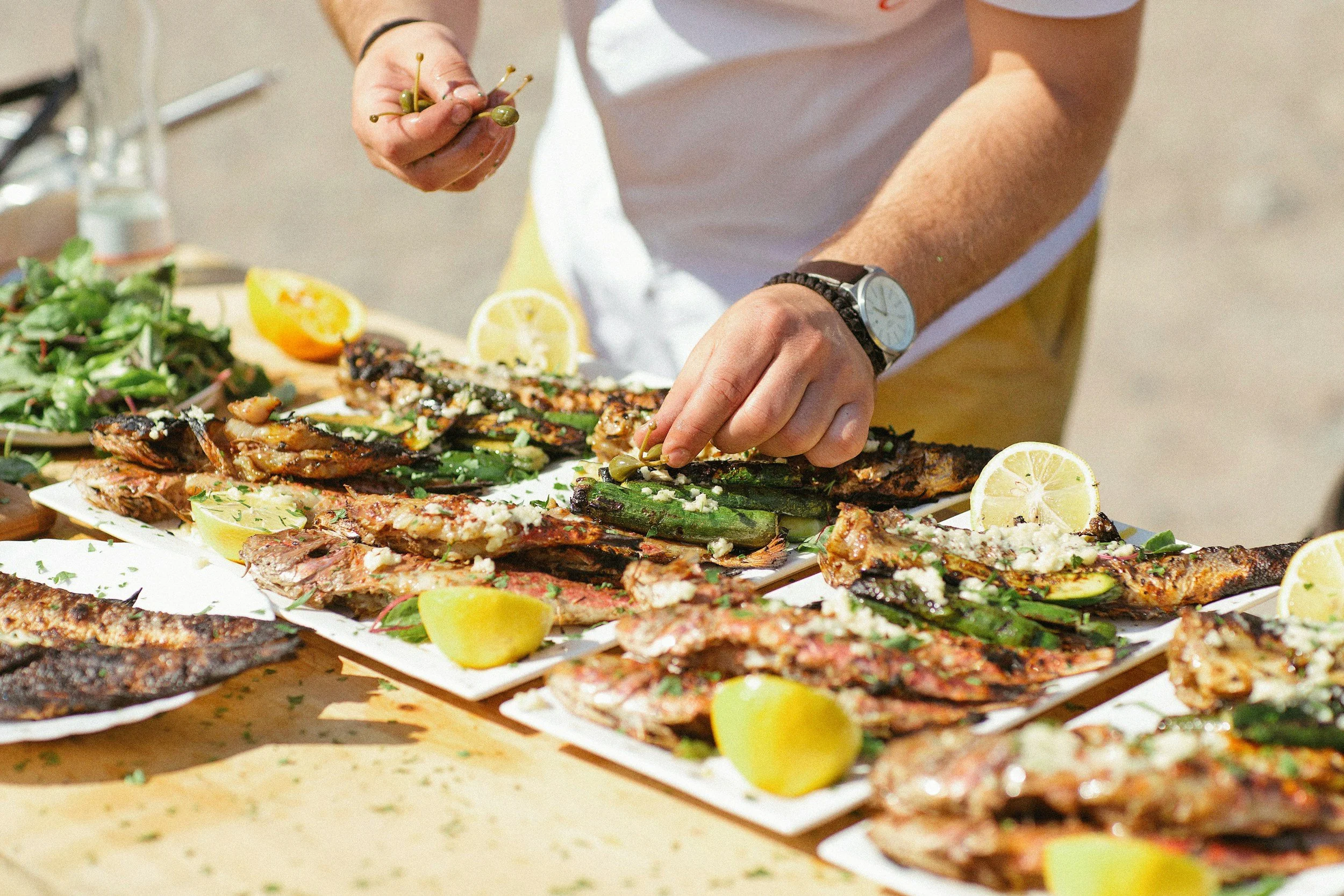 Person garnishing grilled fish with herbs and lemon slices on a table with more grilled fish and lemon wedges.