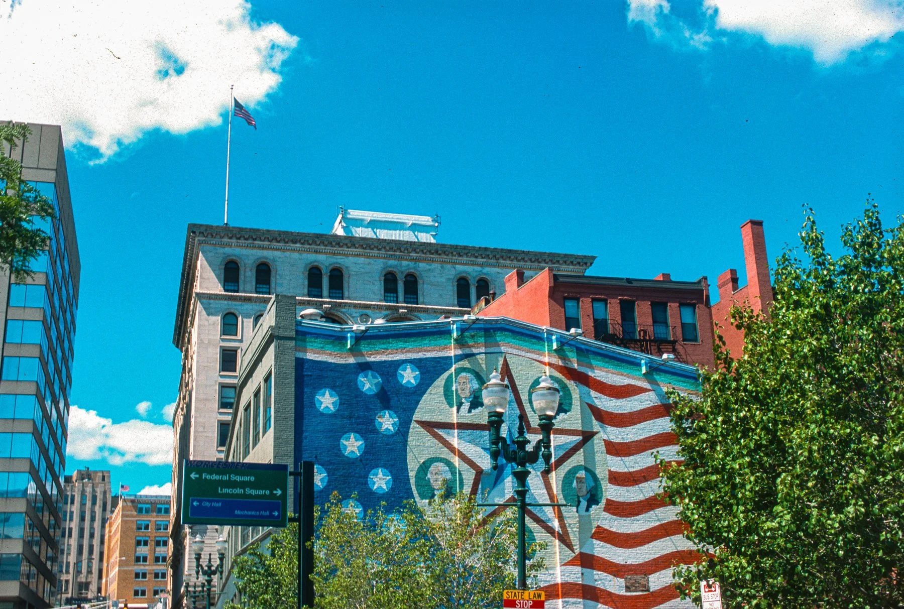 Colorful mural on a building wall featuring a star with images of historic figures, American flag motifs, and a circular emblem, with trees in the foreground and a bright blue sky with clouds.