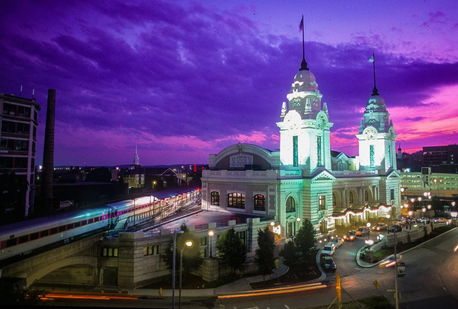 A historic building with two lit towers against a colorful purple and pink sunset sky. A train is passing by on elevated tracks, and there are cars parked along the street below.