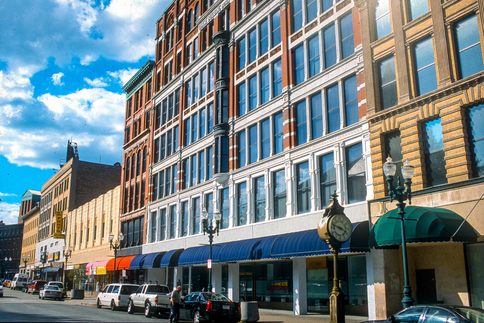 City street with multi-story buildings, parked cars, street lamps, a clock, and a blue awning, under a partly cloudy sky.