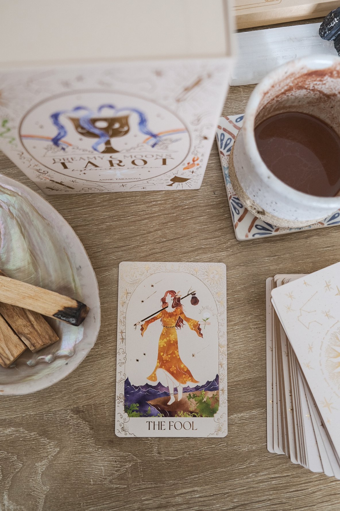 Tarot card titled 'The Fool' depicting a woman in a yellow dress carrying a bag, standing by a mountain and under stars. A cup of hot ceremonial cacao and a tarot deck are also on the wooden table.