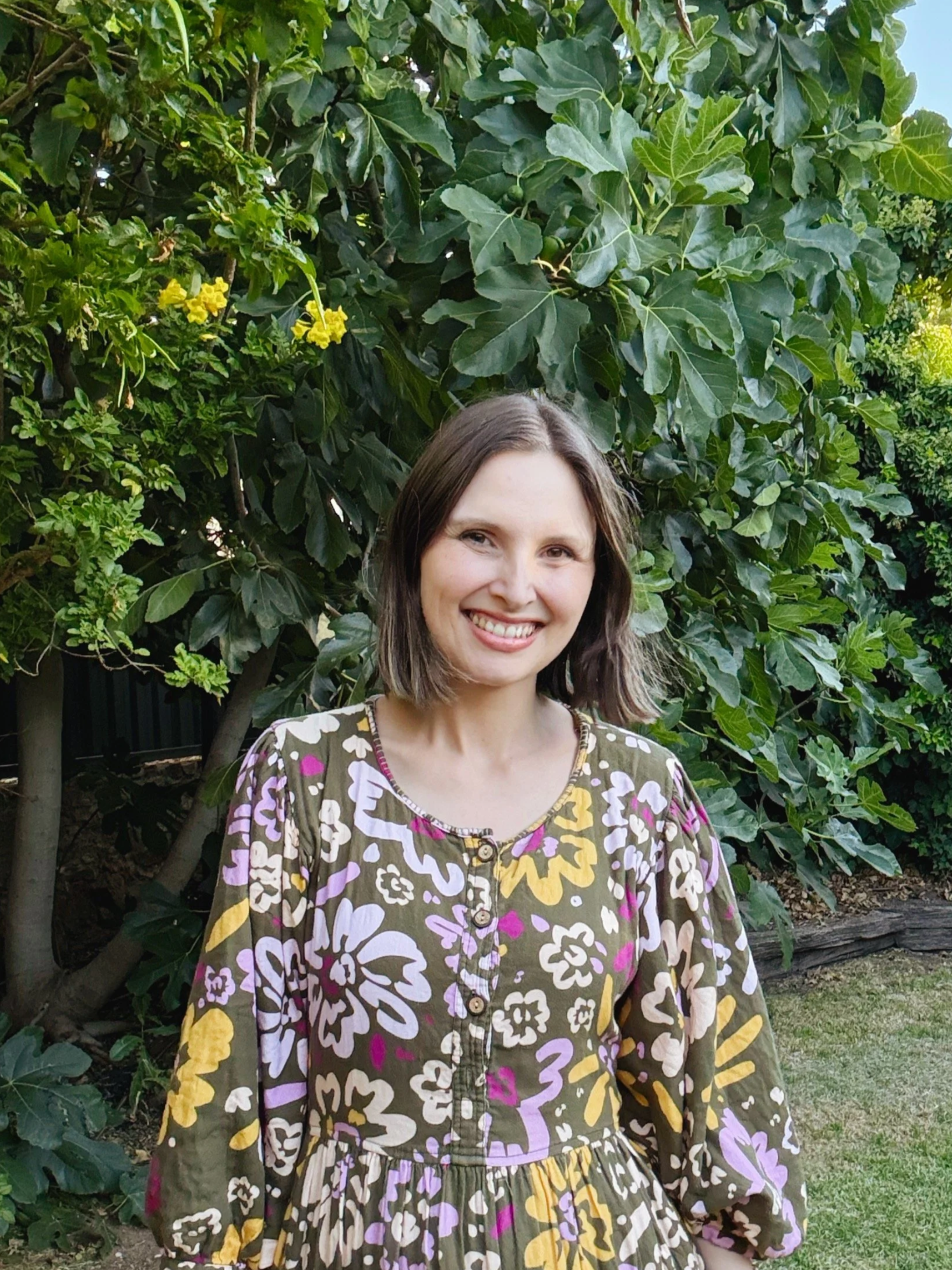 A woman with shoulder-length brown hair smiling outdoors near green bushes with dark green leaves and yellow flowers.