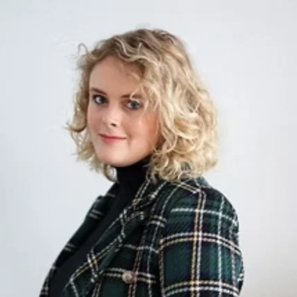 A woman with curly blonde hair wearing a black turtleneck and a green plaid blazer looking at the camera with a slight smile.