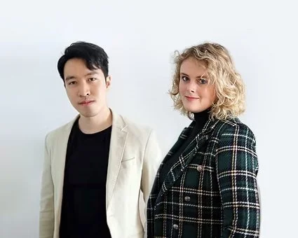 Two people, a man and a woman, standing against a light background, facing the camera. The man has short black hair and is wearing a light-colored blazer over a black shirt. The woman has curly blonde hair and is wearing a dark plaid jacket.