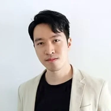 Portrait of a young man with black hair, wearing a black shirt and light-colored blazer, standing against a plain background.