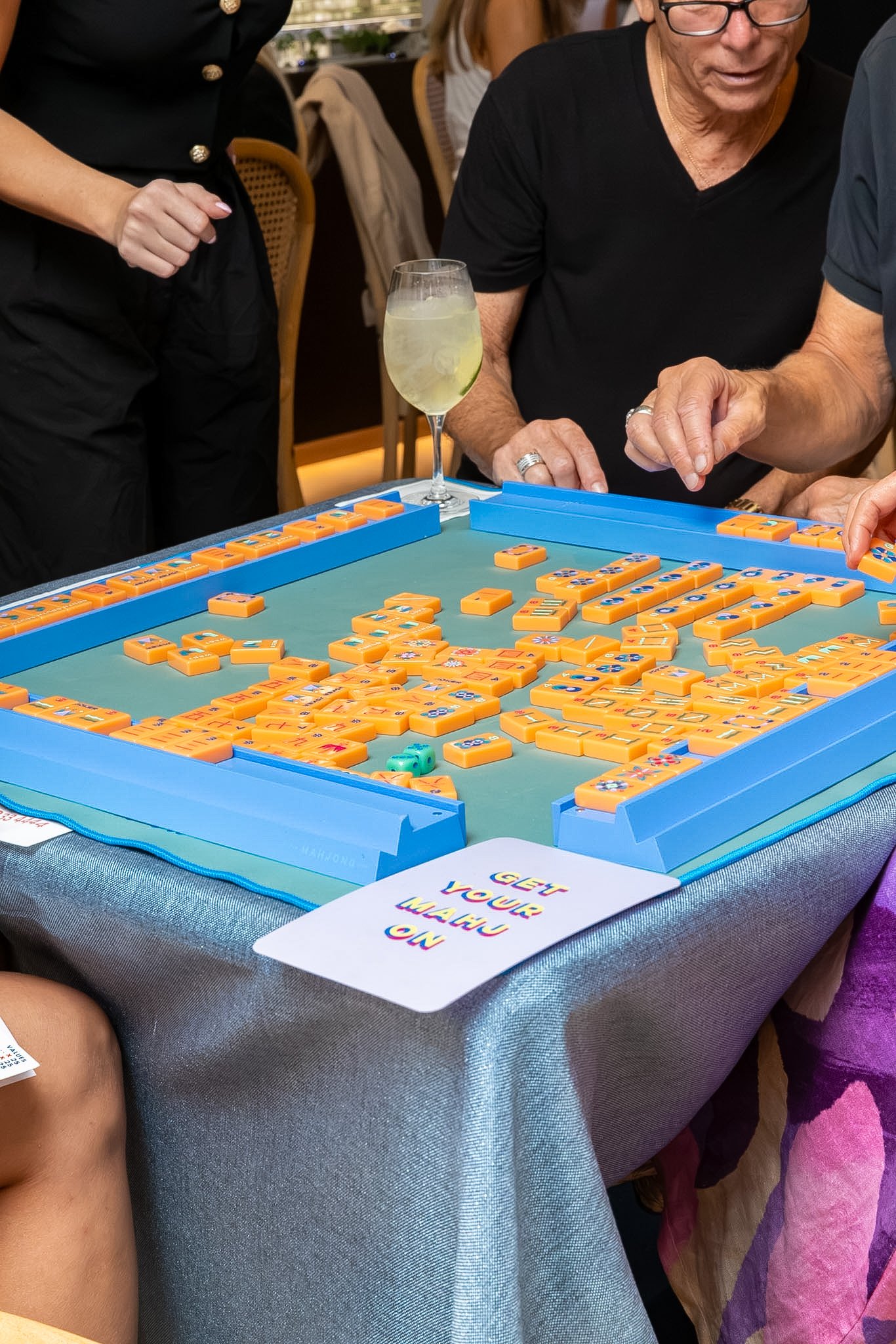 People playing a game of Mahjong at a table in a social setting, with a drink and a card on the table.