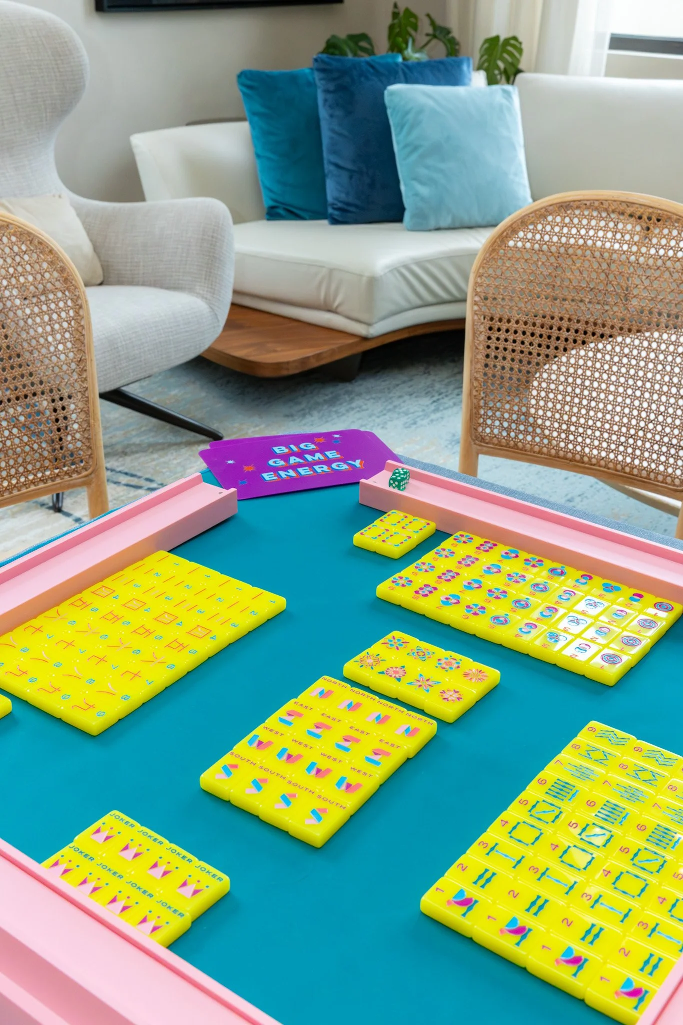 Close-up of a bright teal tiled game board with yellow numbered cards and colorful domino-like tiles, part of a board game setup in a cozy living room with white and blue cushions, wicker chairs, and houseplants in the background.