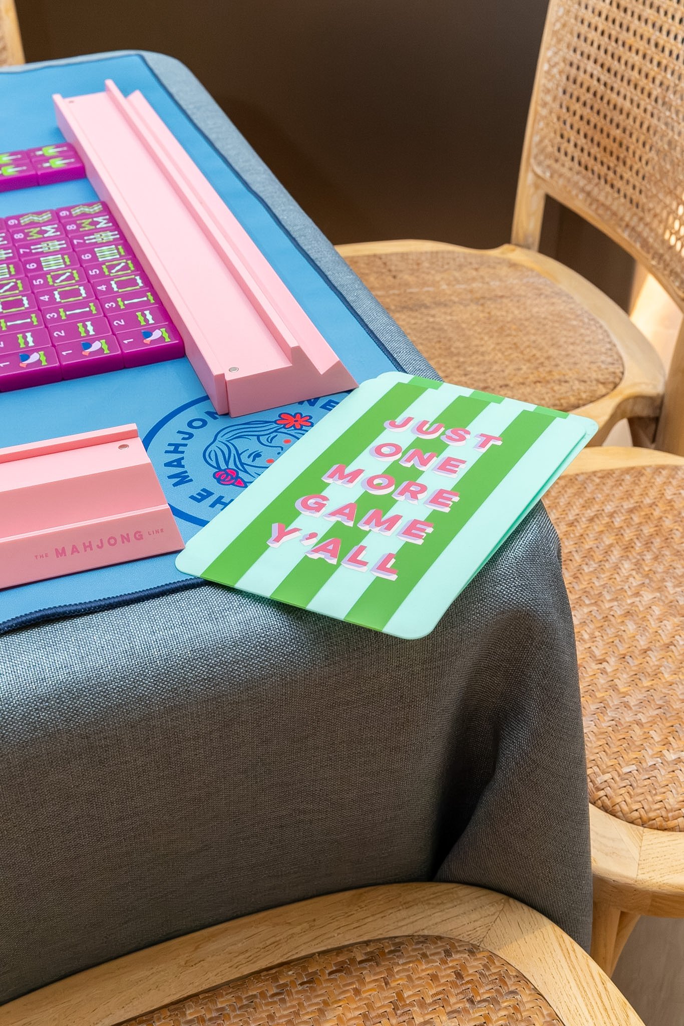 A table decorated with a colorful game board, a pink plastic game piece, and a card with the message 'JUST ONE MORE GAME Y'ALL'. There are wooden chairs with woven seats around the table.