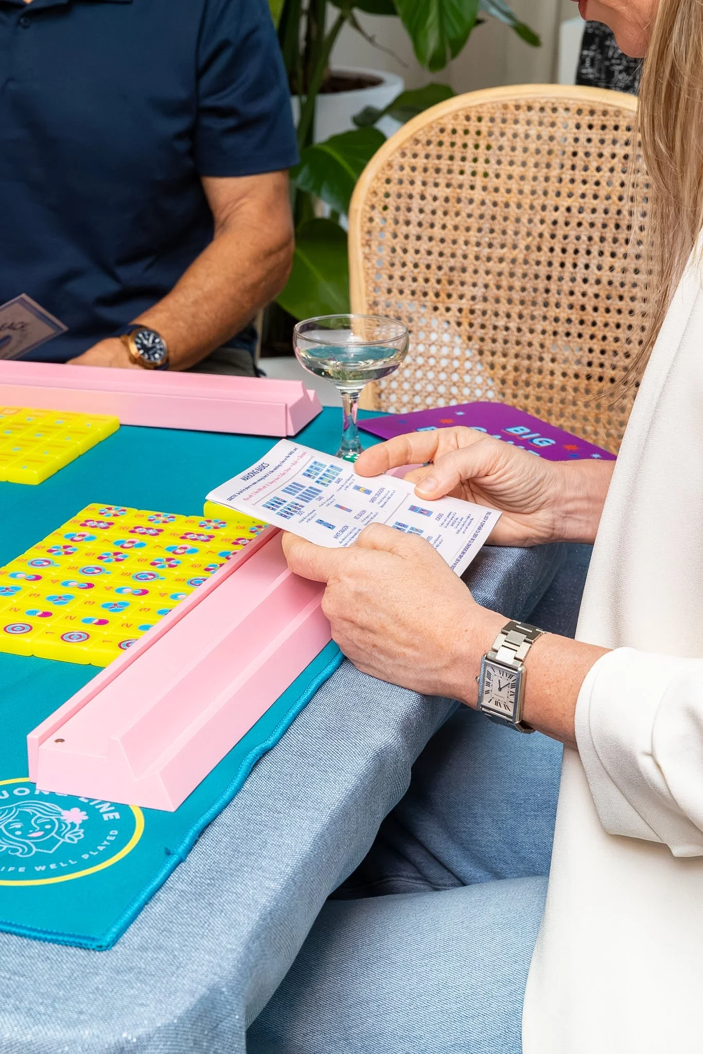 People playing bingo at a table with bingo cards, a pink container, a glass of water, and large green plants in the background.
