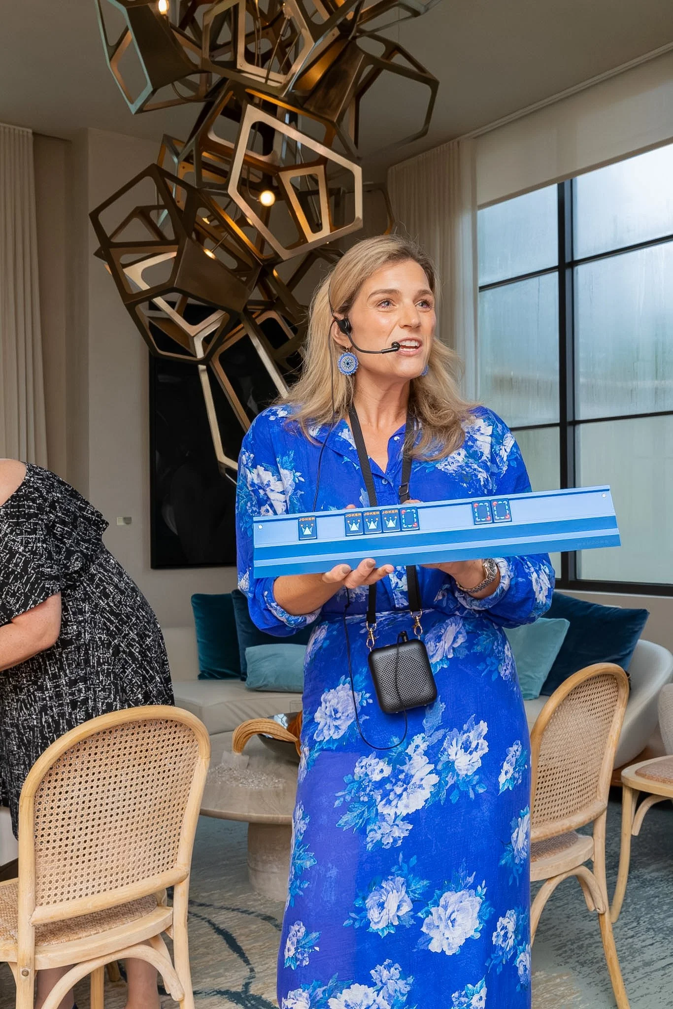 A woman with blonde hair wearing a blue floral dress and a headset, holding a blue board with game symbols.
