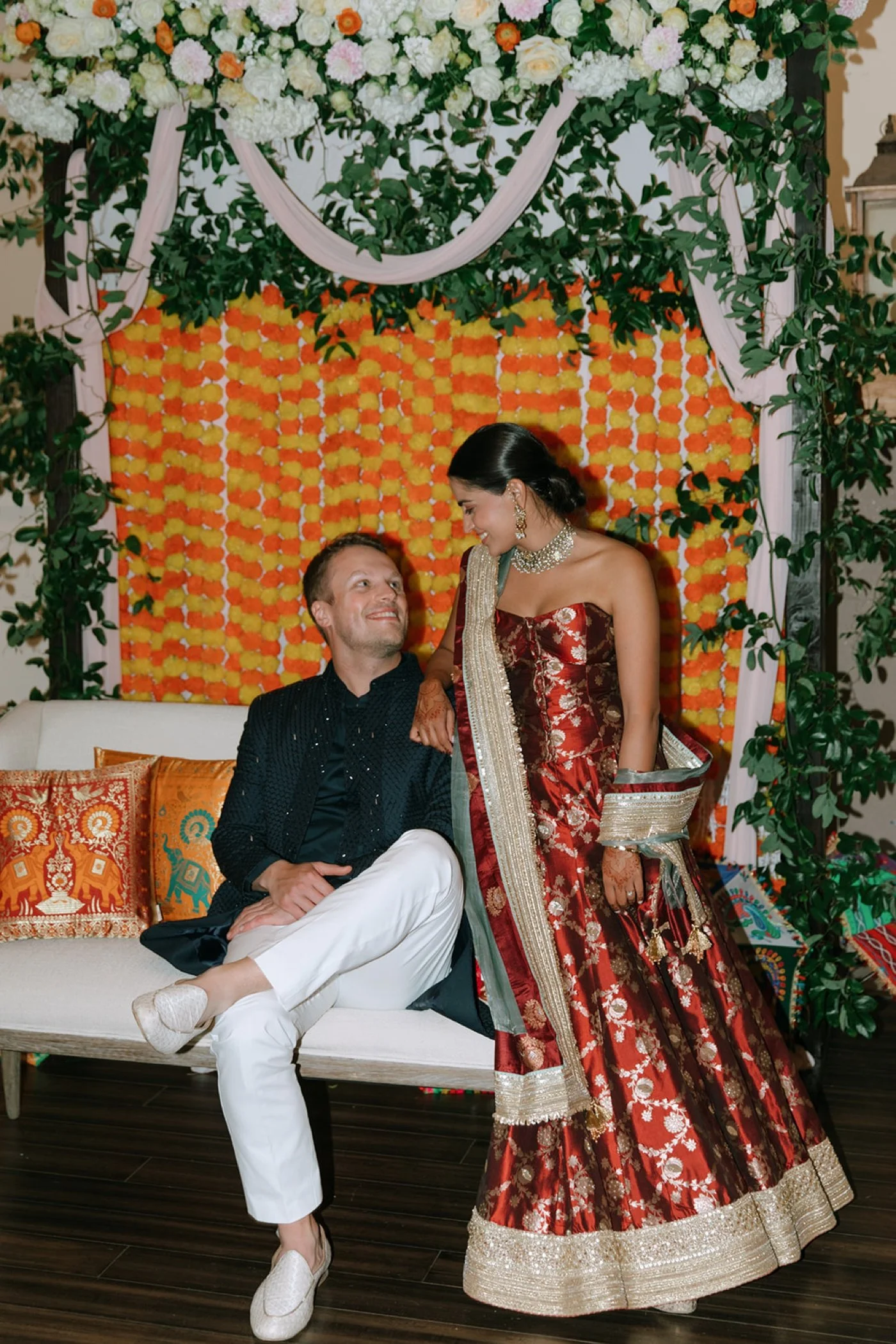 Bride and groom in front of an orange and yellow floral backdrop at their sangeet ceremony