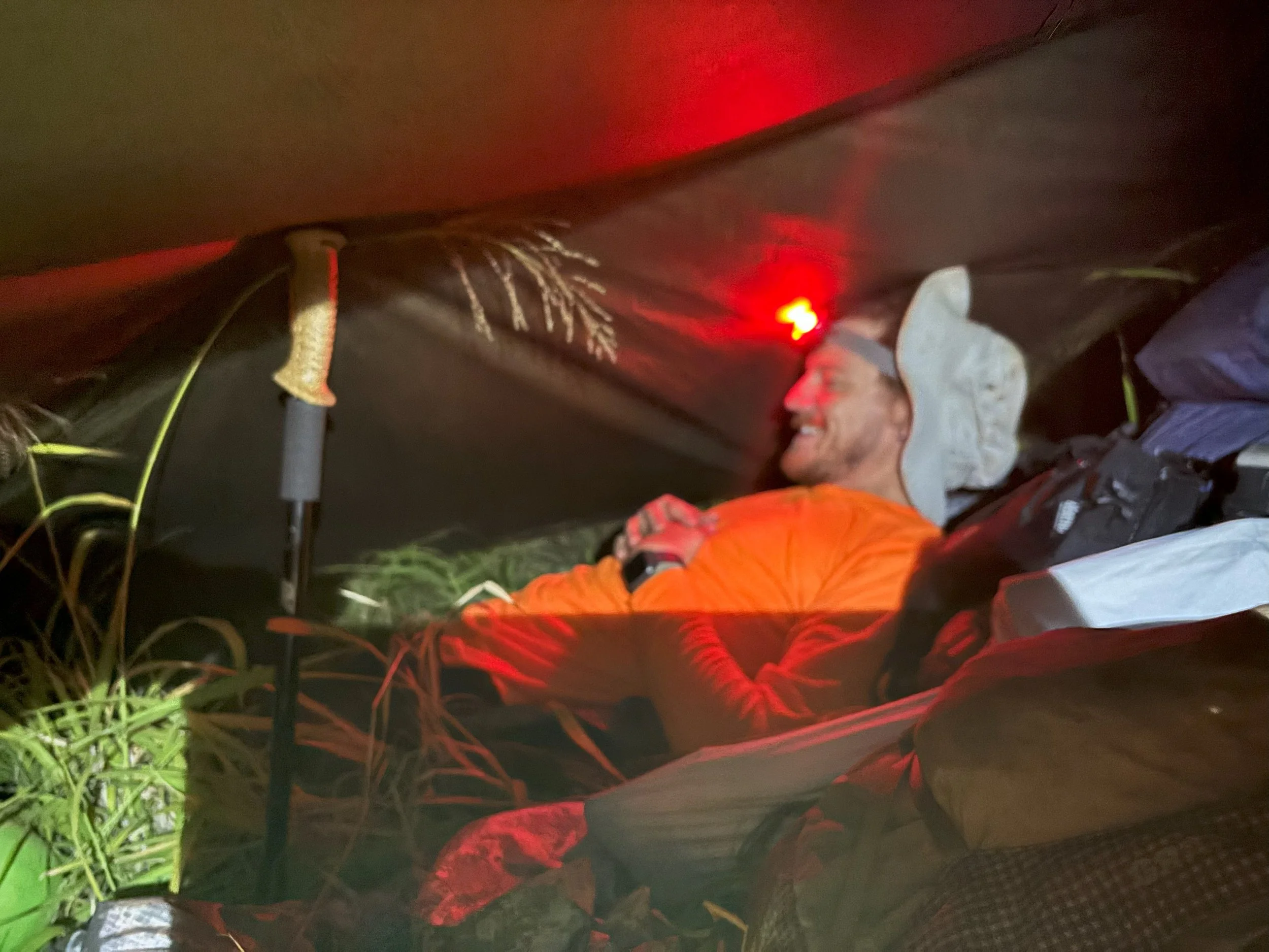 A hunter camping in a tent at night, smiling and wearing an orange jacket and a hooded hat, with a headlamp on their forehead.