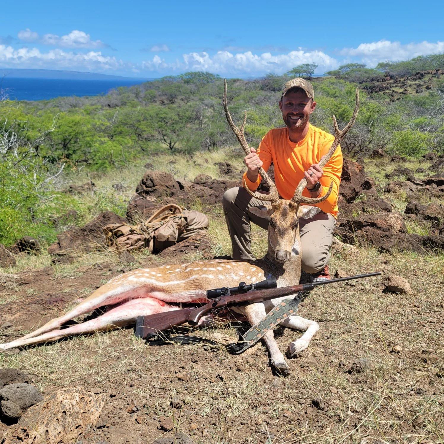 A man in an orange shirt kneeling next to a harvested deer with antlers, holding the antlers, outdoors in a grassy, rocky landscape with trees and a distant ocean under a blue sky.