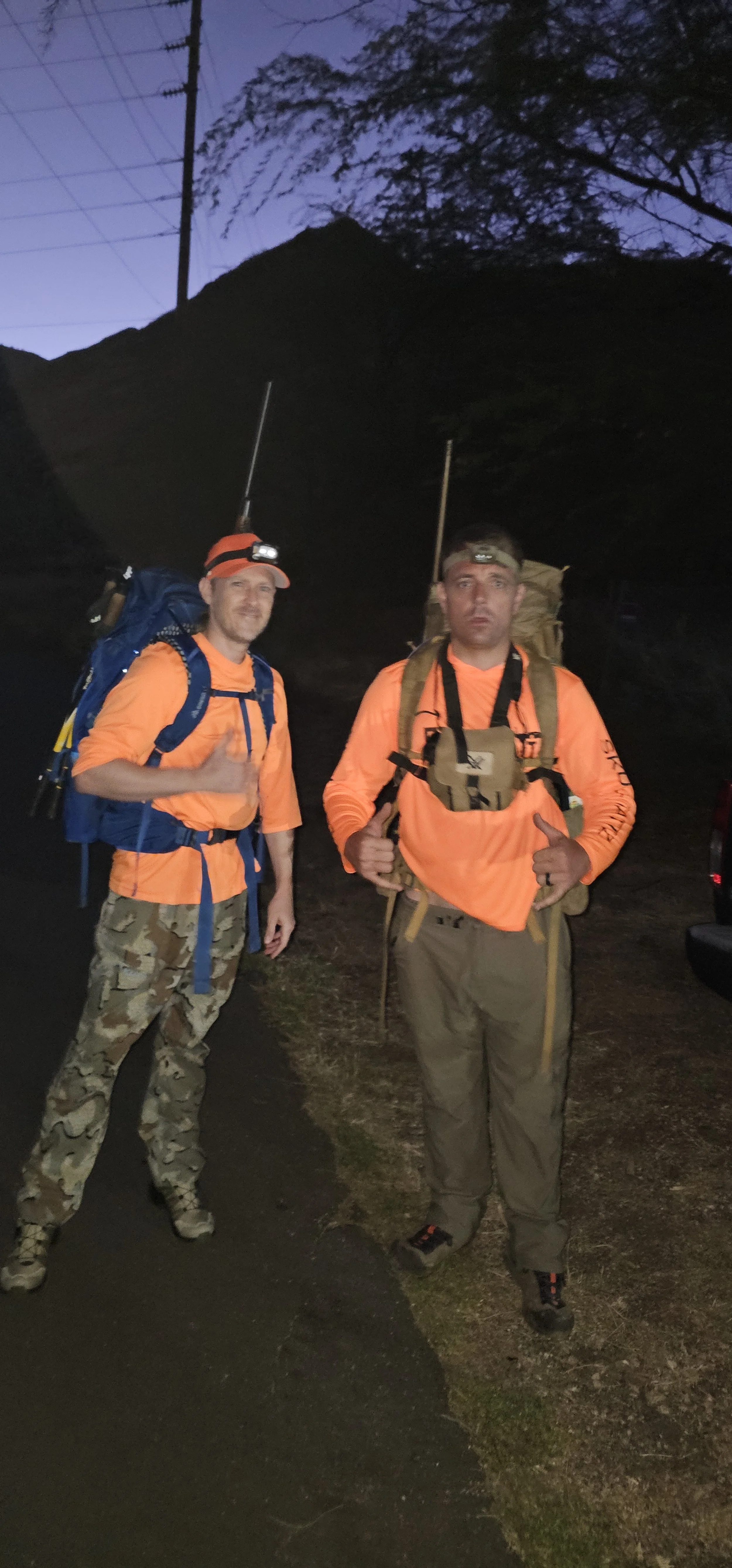 Two male hikers wearing orange shirts, camouflage and tan pants, and headlamps with backpacks, standing outdoors at dusk with a dark sky and silhouette of a mountain and trees in the background.