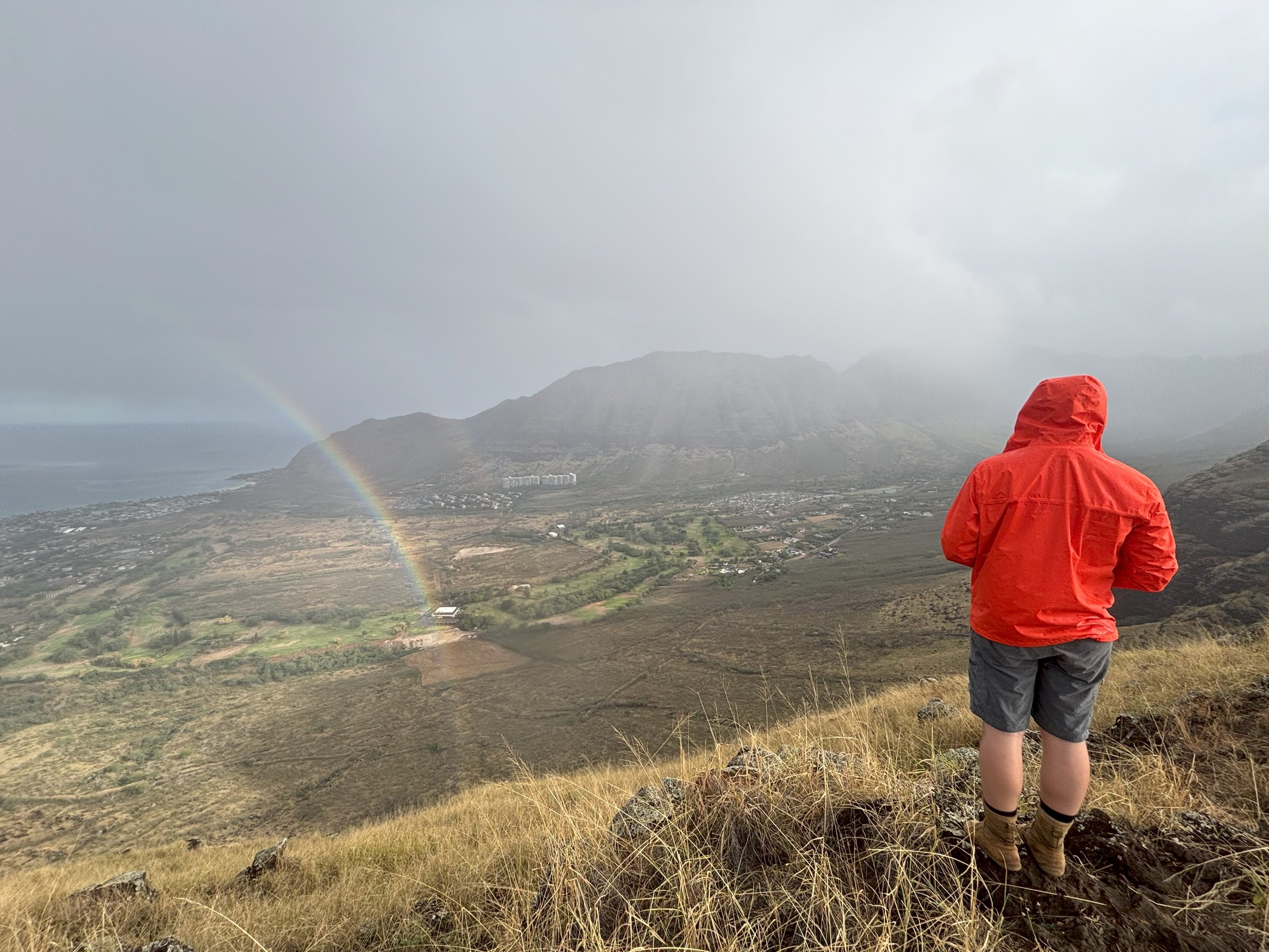 Hunter in a red jacket and shorts standing on a grassy hill, overlooking a valley with a rainbow and mountainous landscape in the distance, partly cloudy sky.