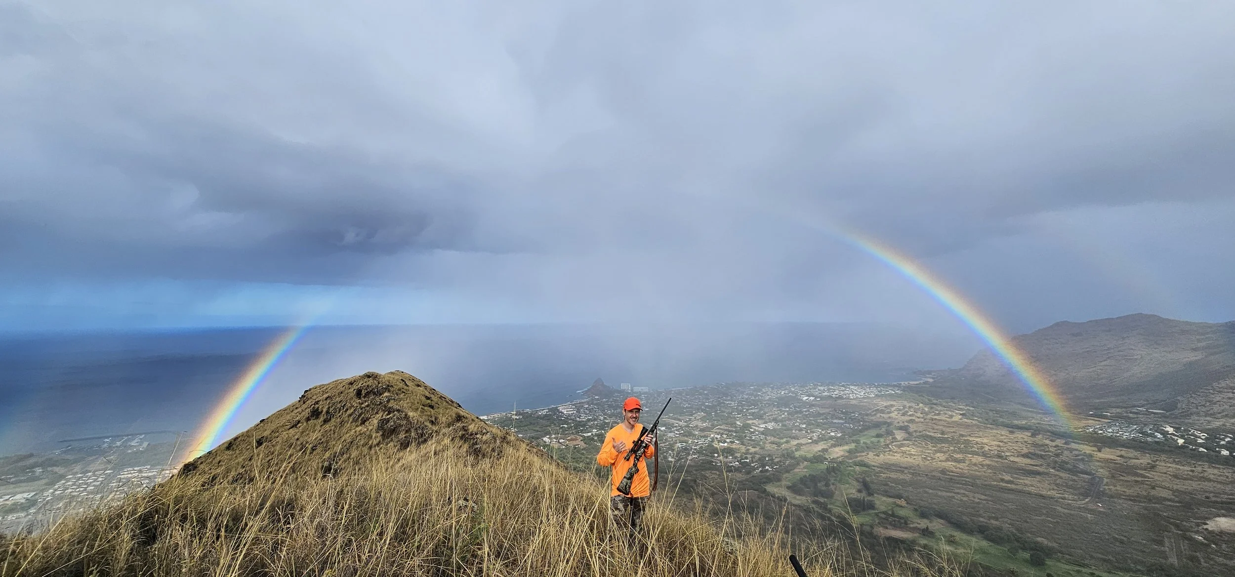 A person in orange outdoor clothing holding a rifle on a grassy mountain ridge with a double rainbow in the cloudy sky and a view of a valley and ocean below.