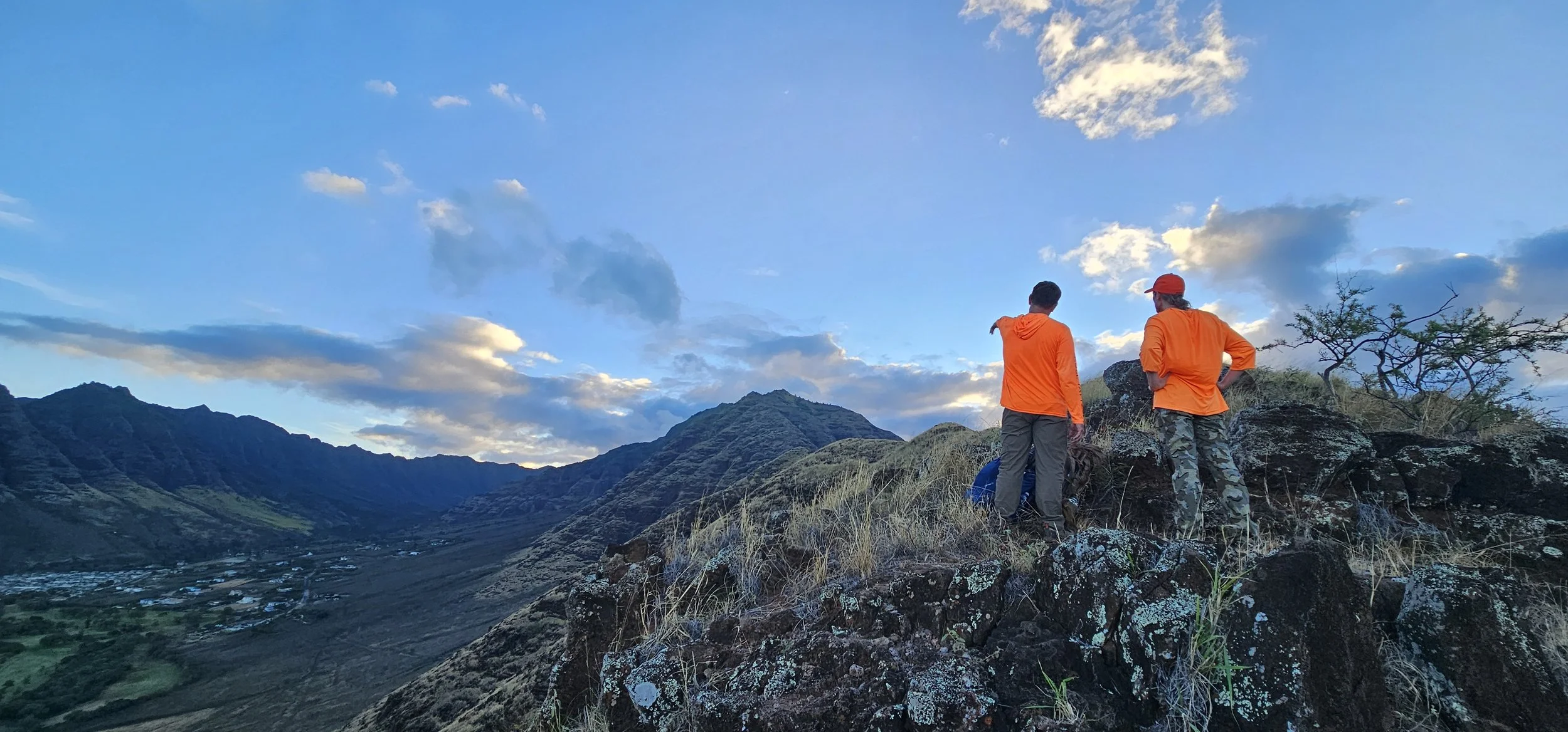 Two men in orange jackets standing on a rocky hillside, overlooking a valley with mountains in the background, under a partly cloudy sky.