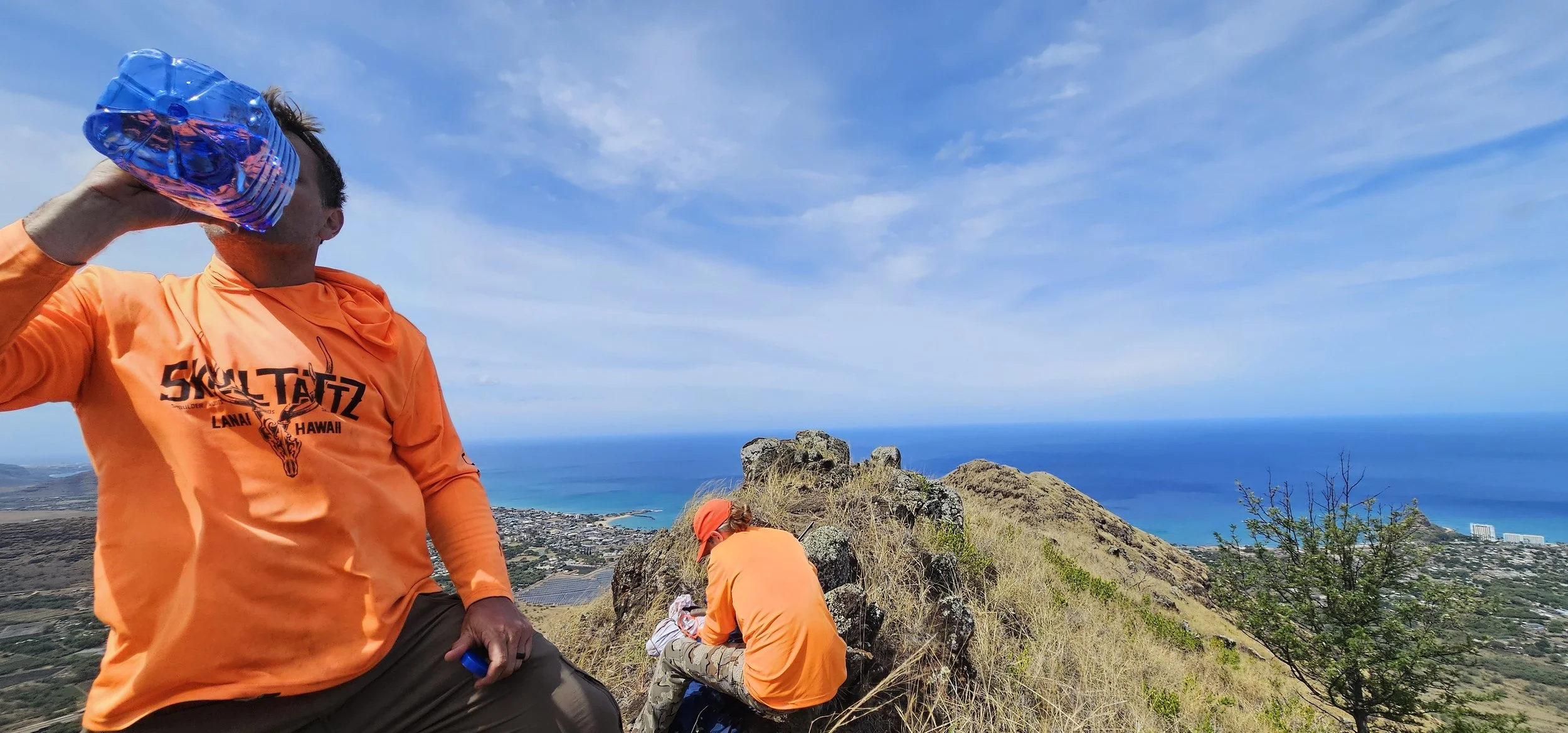Two people in orange shirts on a hillside with a view of the ocean and a town below, one drinking water and the other sitting.