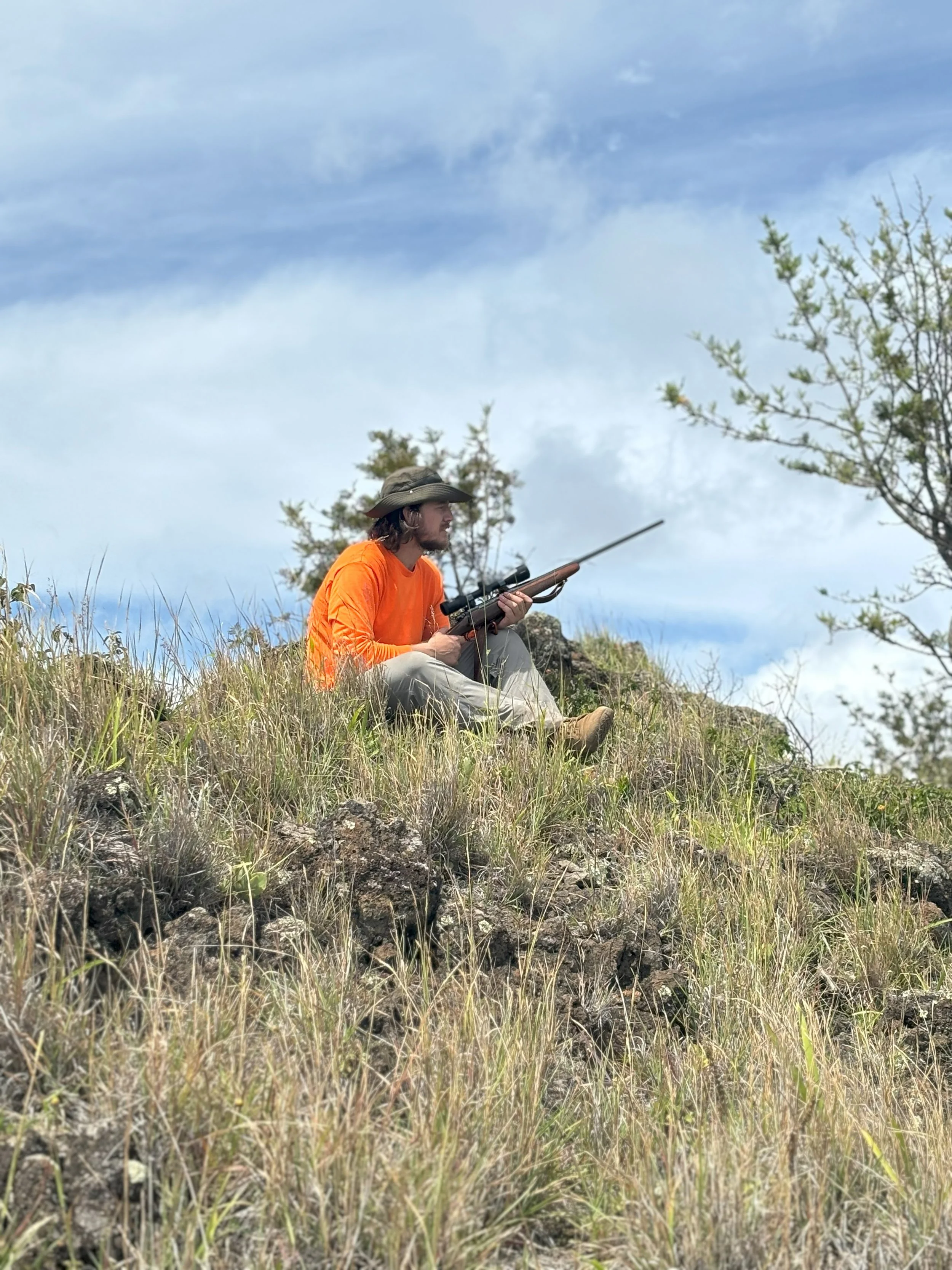 Man sitting on grassy hillside with a rifle, wearing an orange shirt and a wide-brimmed hat, surrounded by small trees and rocks, under a partly cloudy sky.