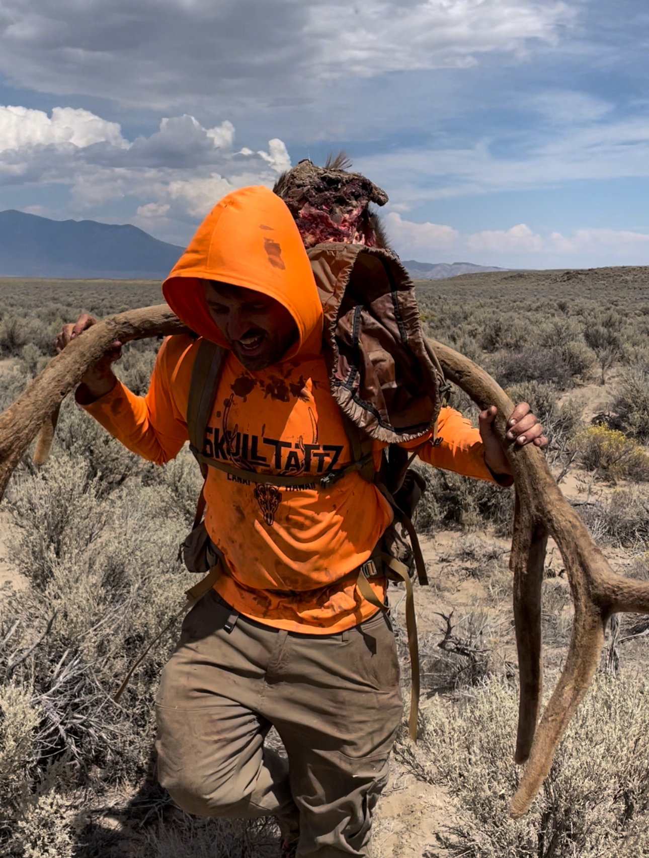 A person wearing an orange hoodie and tan pants carrying a large, four-legged skeleton on their shoulders in a desert landscape with sagebrush and mountains in the background.