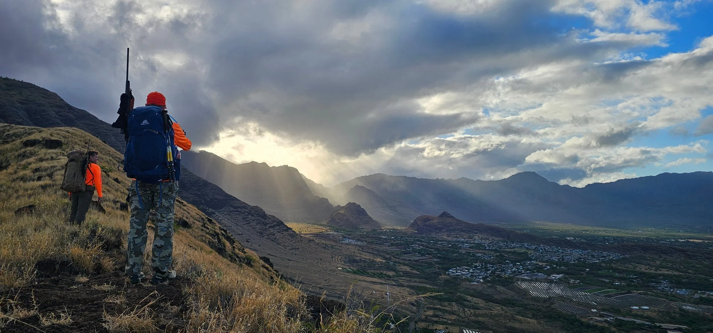 Two hunters with hunting packs and rifles standing on a grassy mountain ridgeline in Hawaii, overlooking a valley with fields and mountains in the distance, under a cloudy sky with rays of sunlight breaking through.
