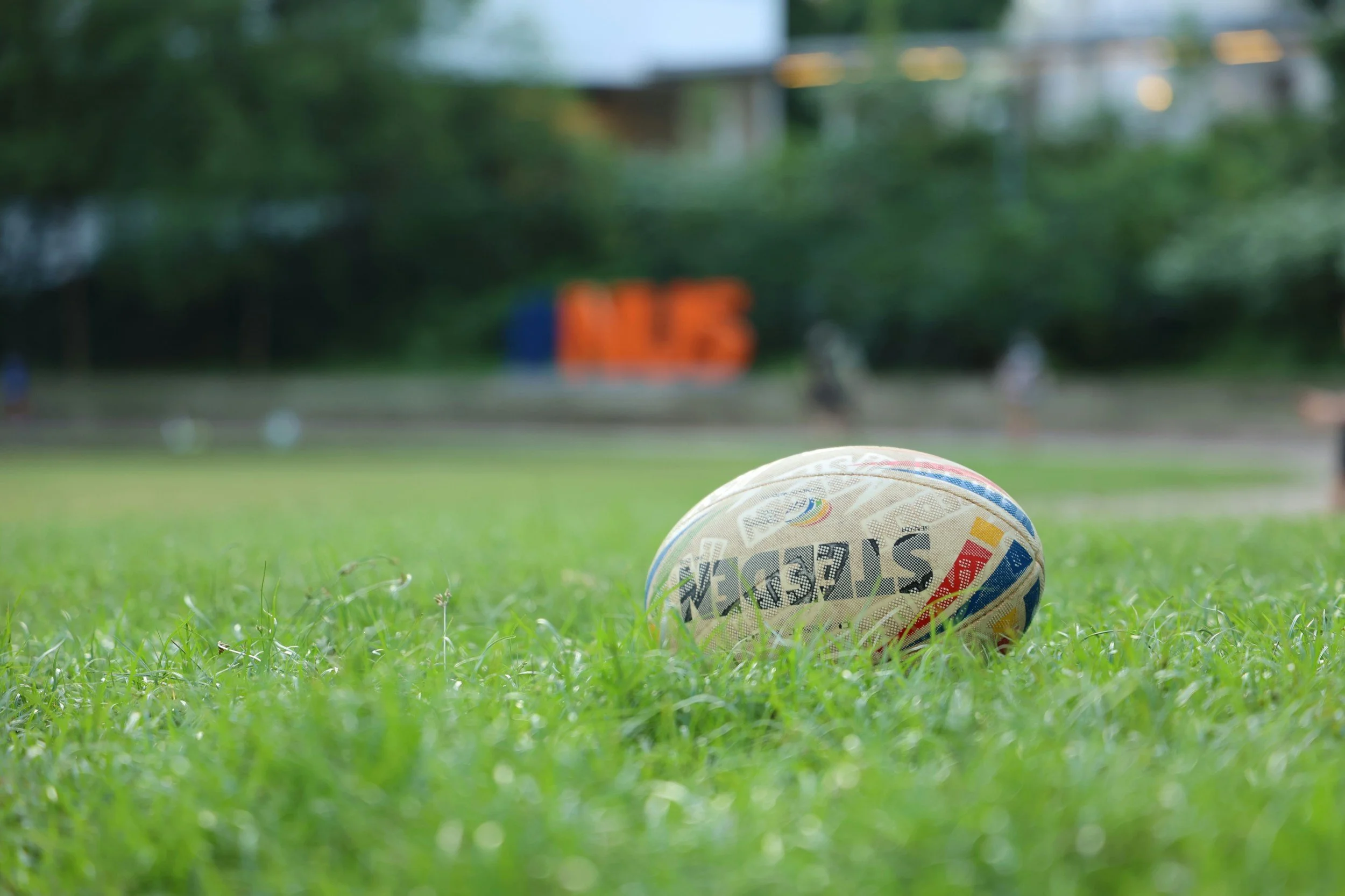 A rugby ball on a grassy field with a blurred background of trees and a large sign that reads 'NIES' in orange letters.