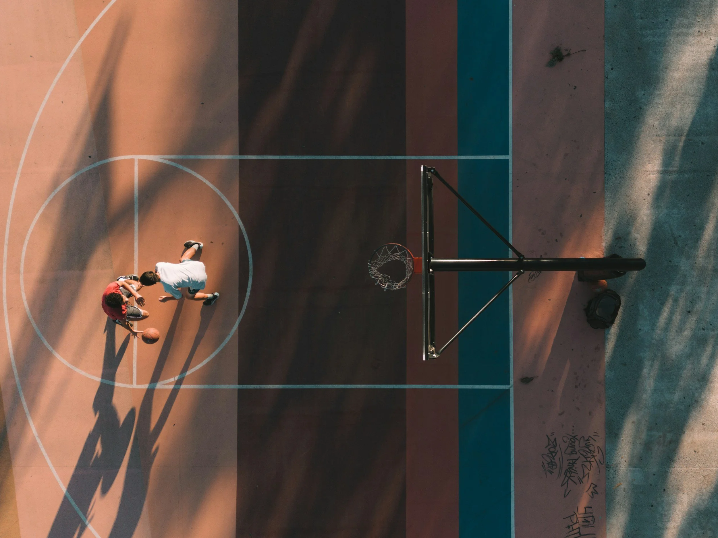Two people playing basketball on an outdoor court, seen from above. The court has markings and a basketball hoop. The players are near the basket, one with a basketball.