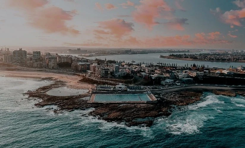 Aerial view of a coastal city at sunset with a beach, buildings, and a body of water.