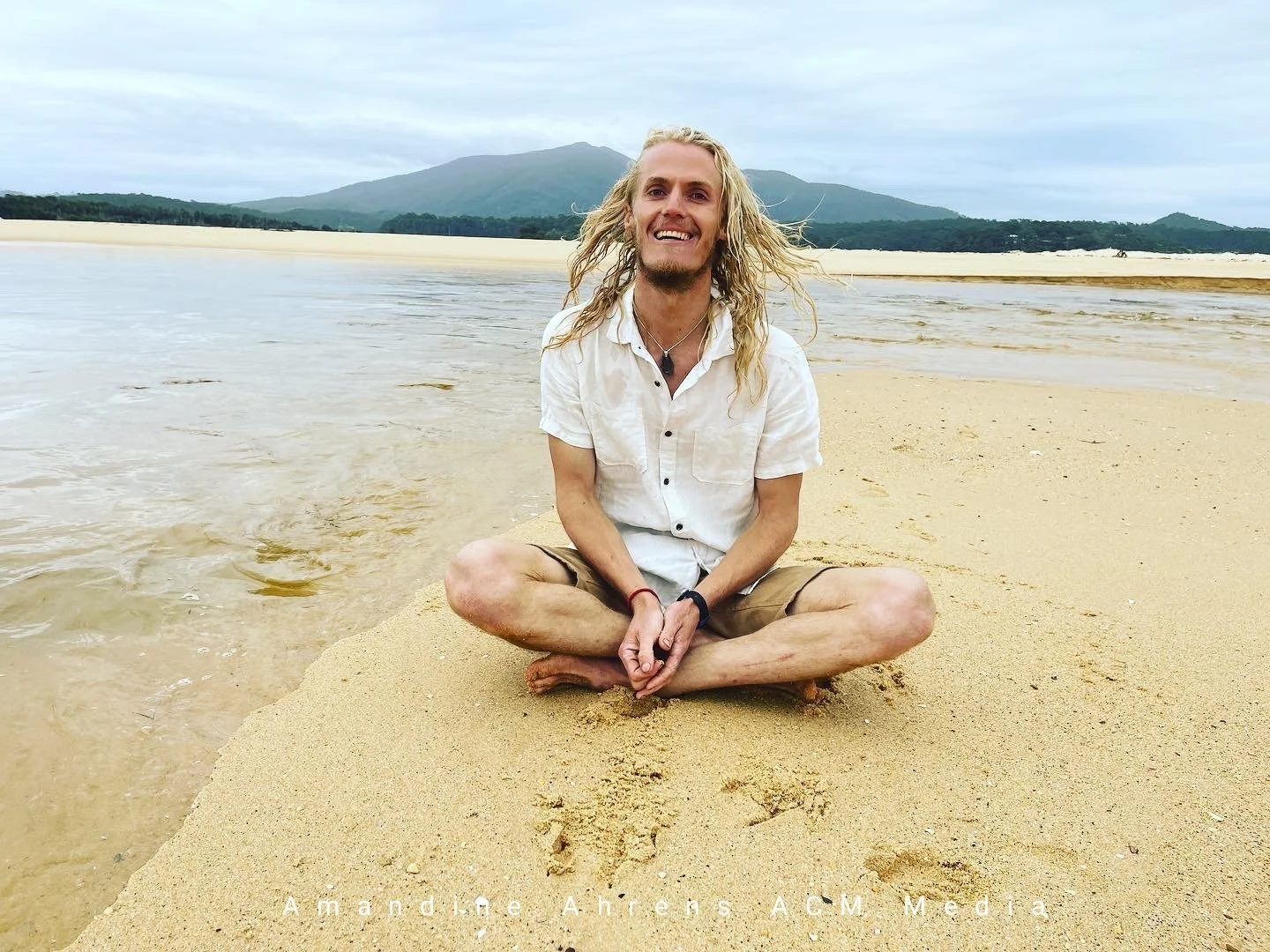 A man with long blonde hair, wearing a white shirt and tan shorts, sits cross-legged on a sandy beach near shallow water, smiling at the camera with mountains and cloudy sky in the background.