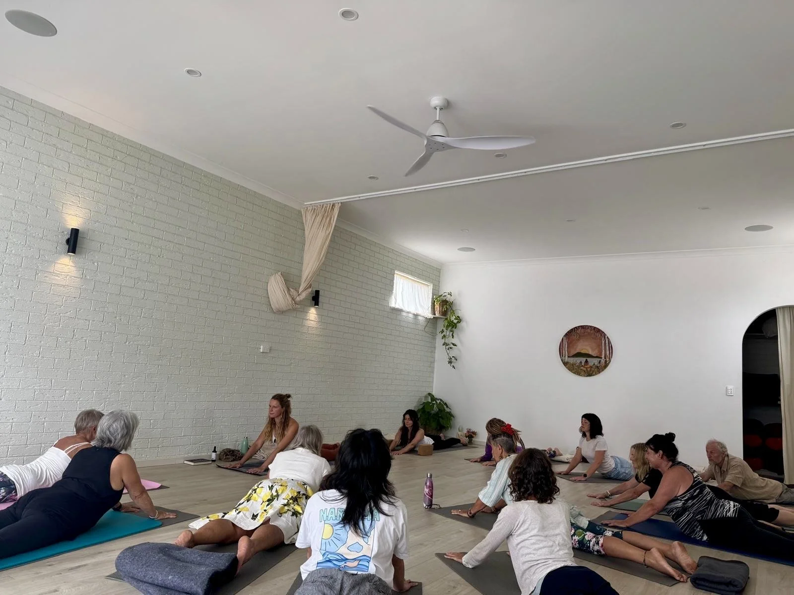 People participating in a yoga or meditation class in a bright, spacious studio with white walls, wooden floors, and a ceiling fan.