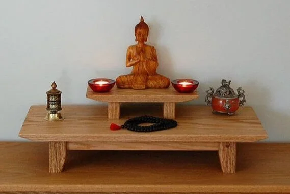 Wooden altar with Buddha statue, two red candles, a small brass lantern, and a decorative bowl with congos on a white wall background.