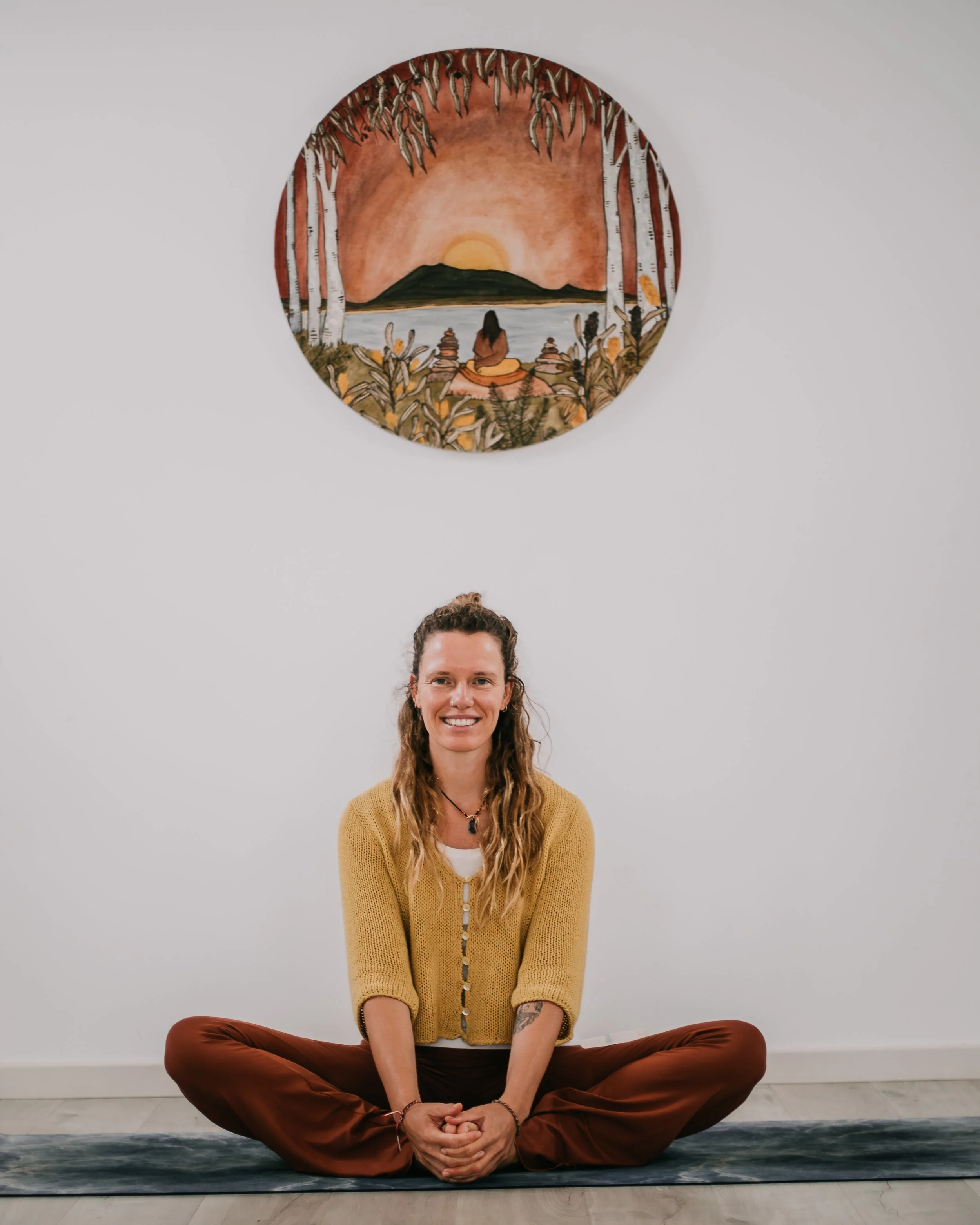 A woman practicing yoga seated on a mat in front of a white wall with a round painting of a sunset landscape with trees, water, and hills.