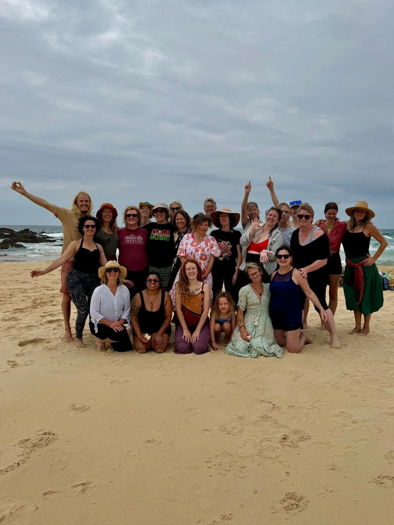 Group of people on the beach posing for a photo, some smiling and making gestures, under cloudy sky.